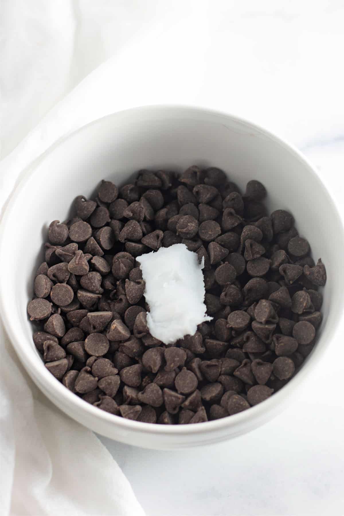 A white bowl filled with dark chocolate chips and a piece of coconut oil in the center.