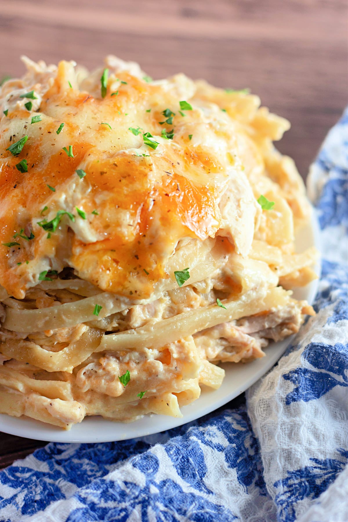 Close-up of a cheesy pasta dish with herbs on top, served on a white plate, next to a blue-patterned cloth.