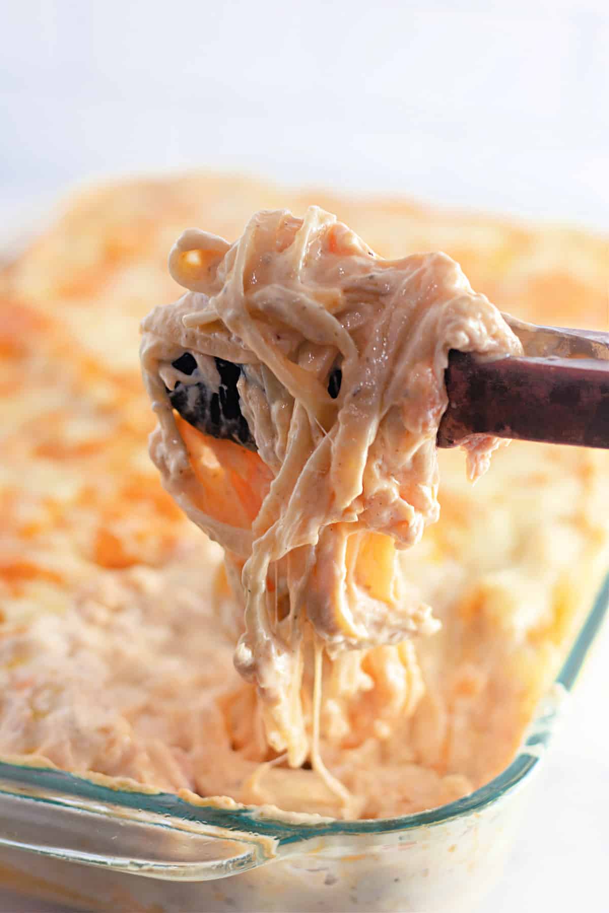 Spoon lifting cheesy pasta from a baked dish in a glass pan.