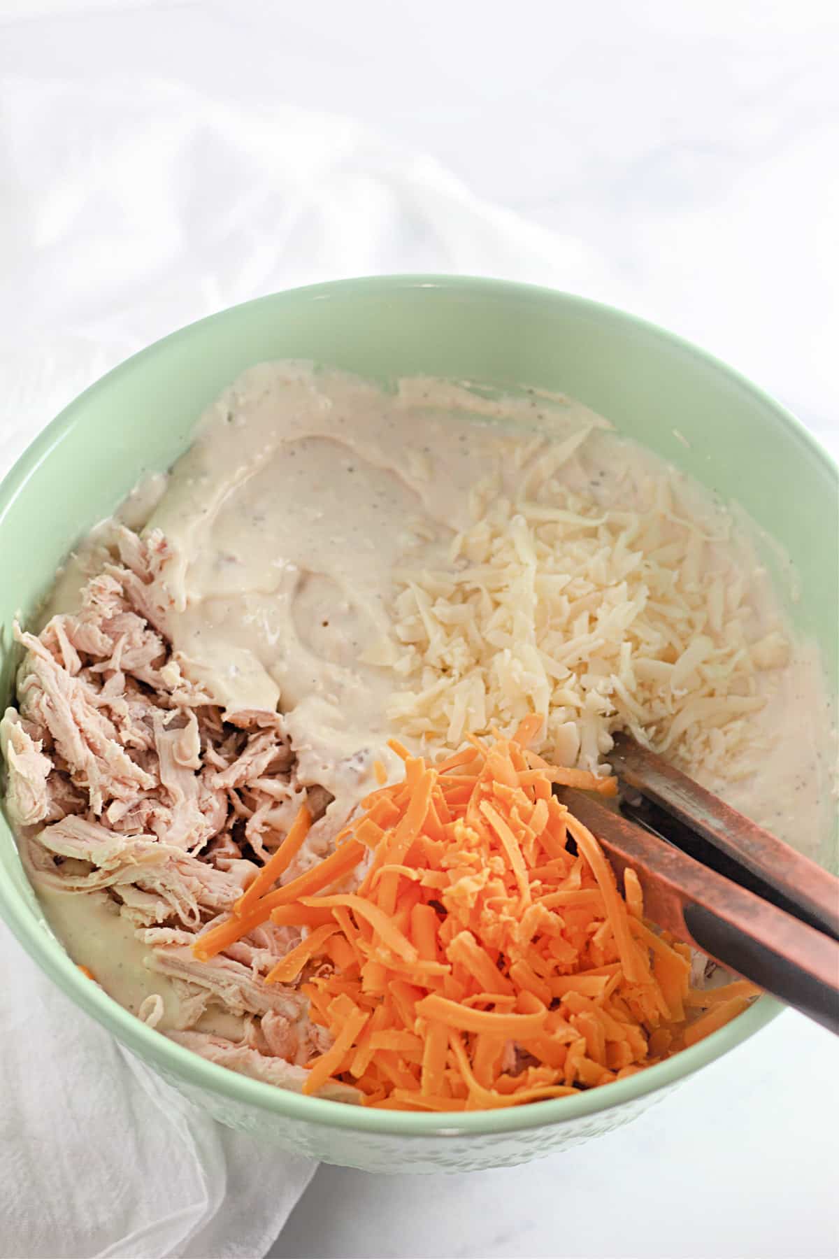 Green bowl with shredded chicken, cheddar cheese, and white sauce being mixed with wooden tongs on a white surface.