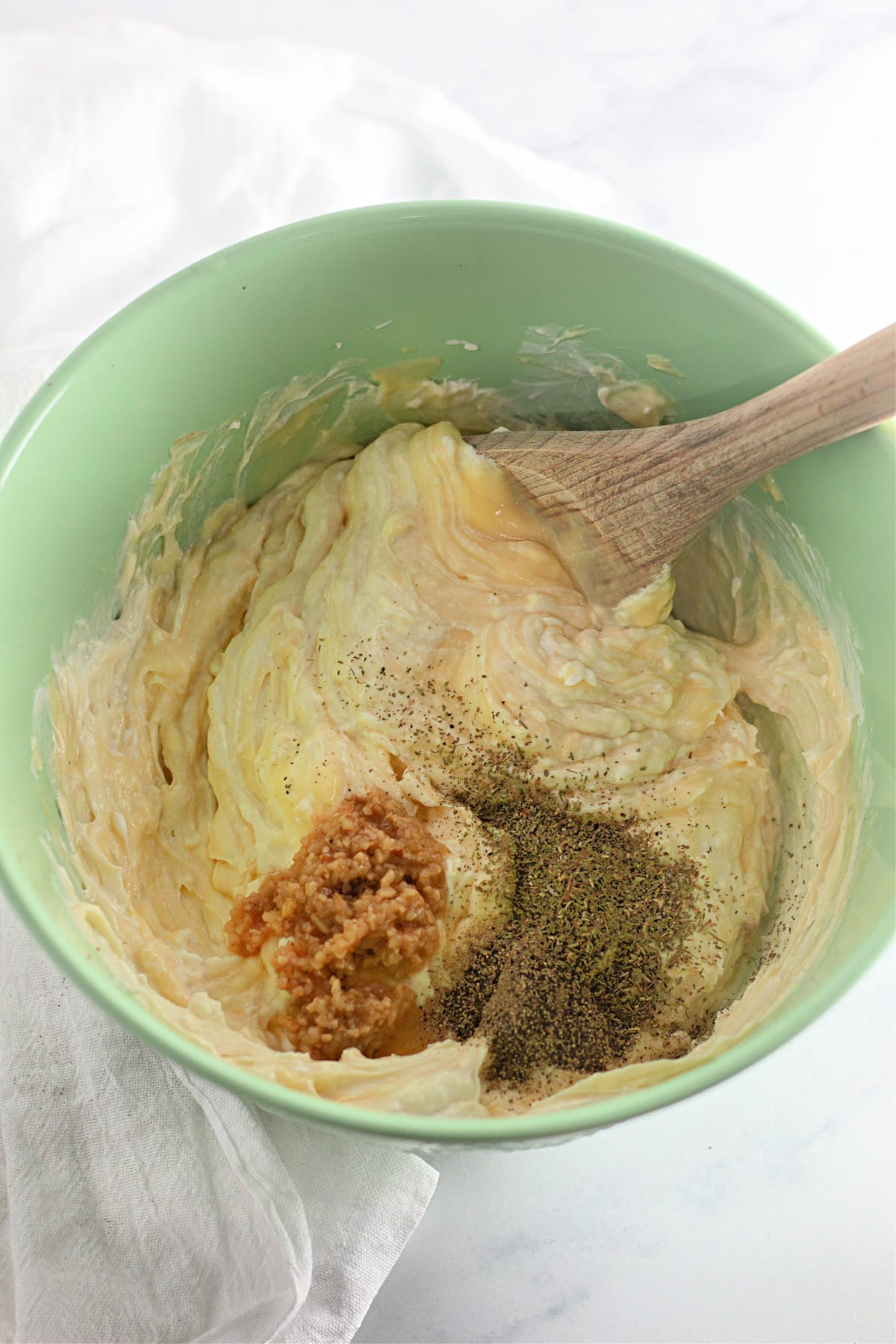 Green bowl with creamy mixture, black pepper, and brown seasoning, stirred by a wooden spoon on a white cloth.