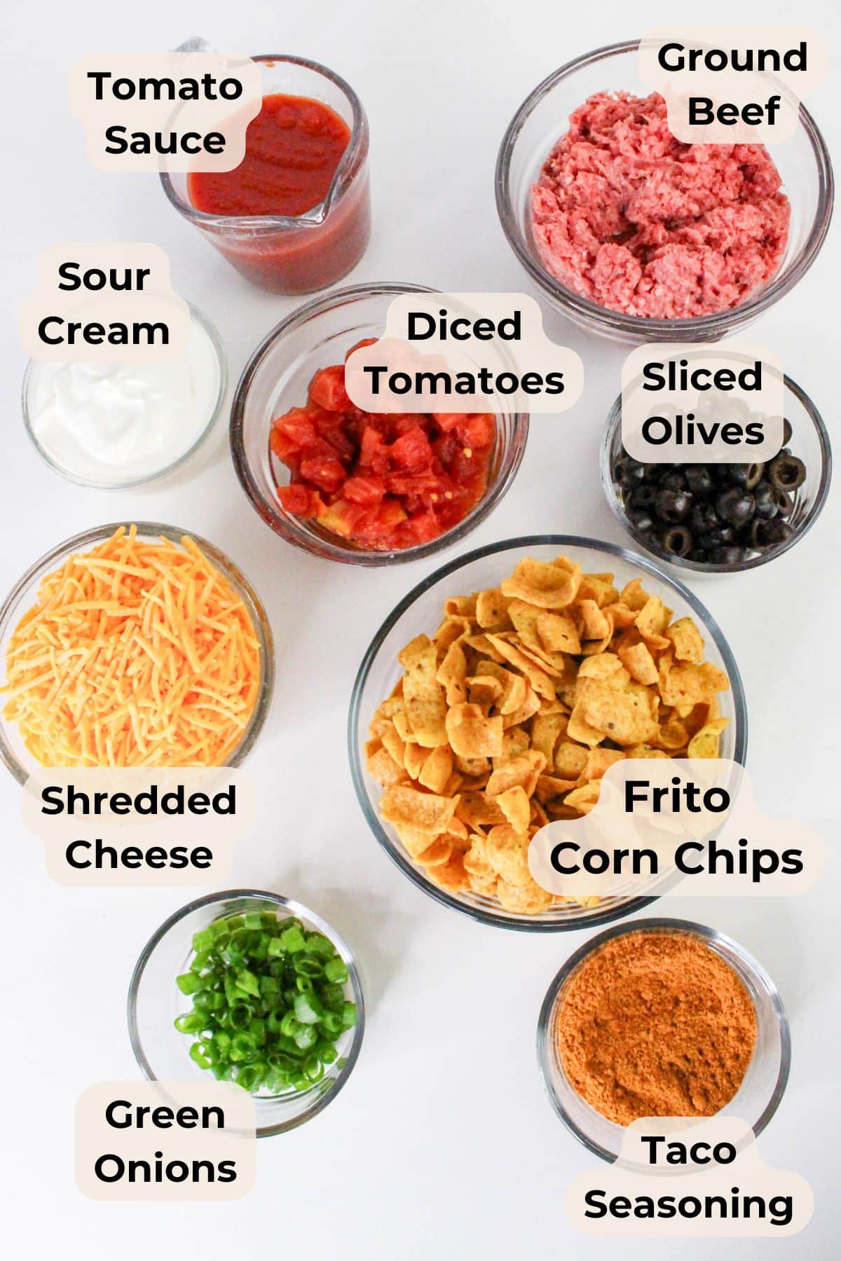 Bowls filled for taco casserole with Fritos, ground beef, tomato sauce, olives, sour cream, cheese, onions, and zesty seasoning.