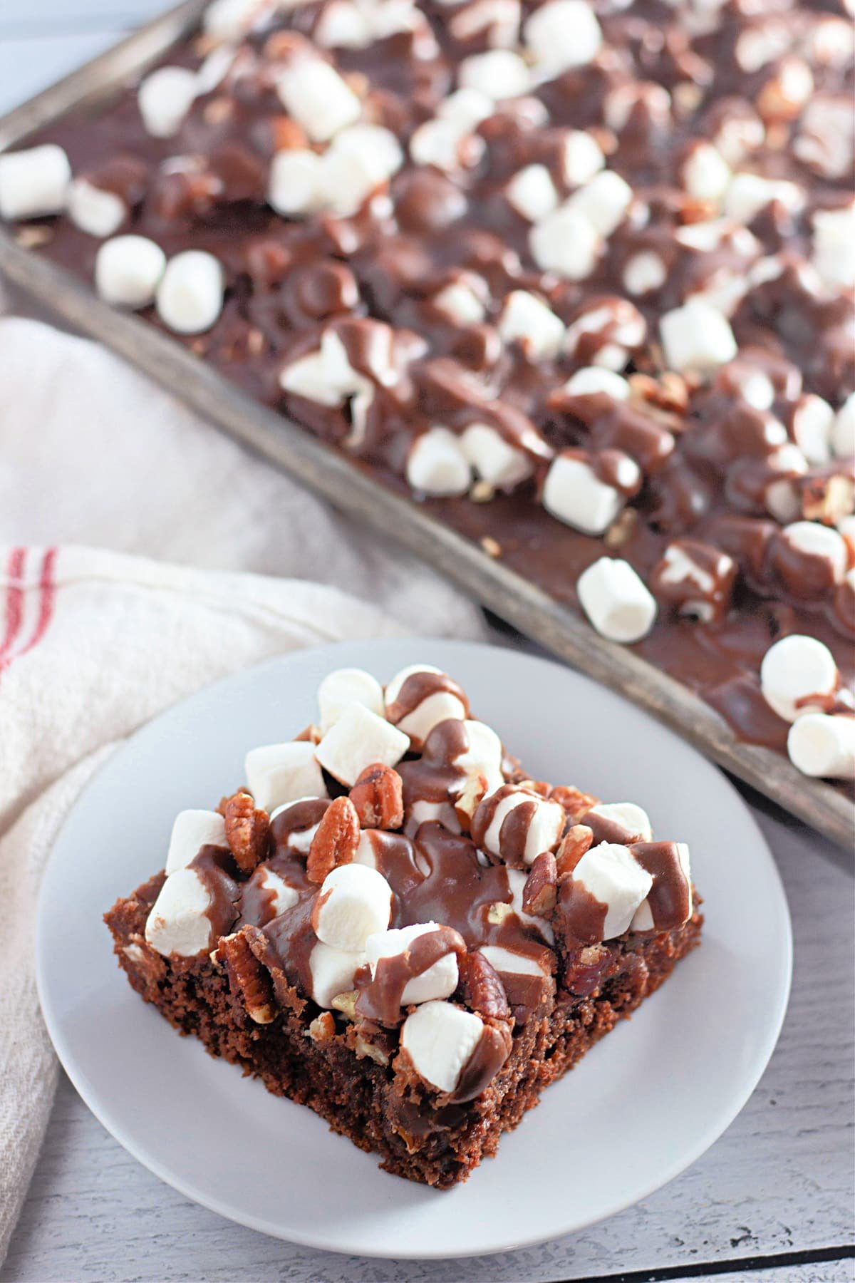 A chocolate dessert bar topped with marshmallows and nuts on a white plate, with more bars on a tray in the background.