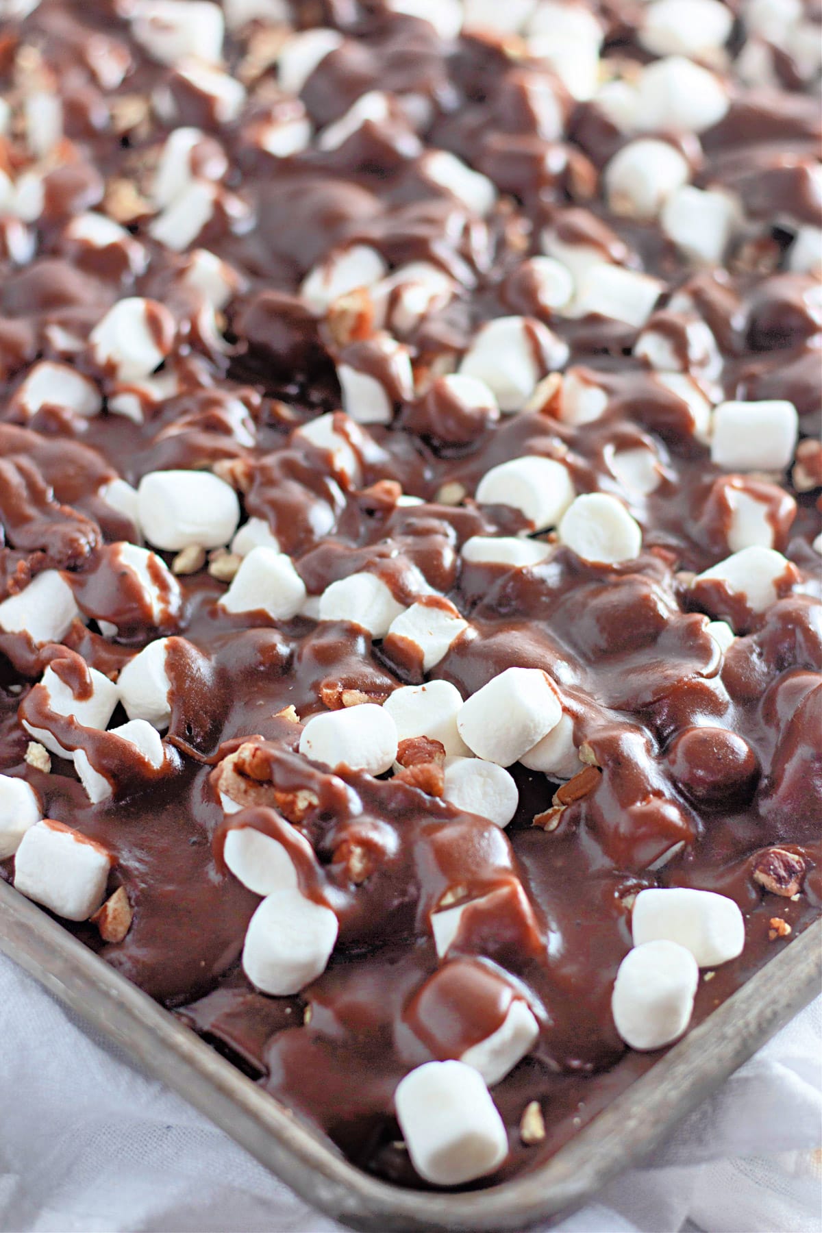 Close-up of a dessert topped with melted chocolate, mini marshmallows, and chopped nuts on a tray.