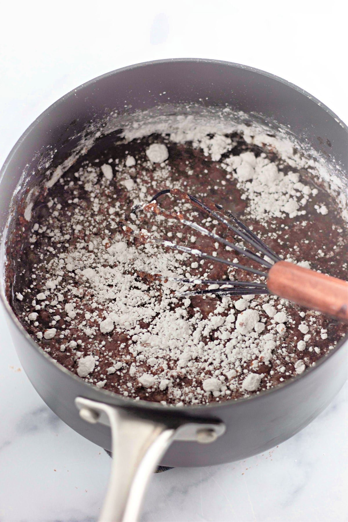 A pot with chocolate batter being mixed, dusted with flour, and a whisk resting inside on a marble surface.