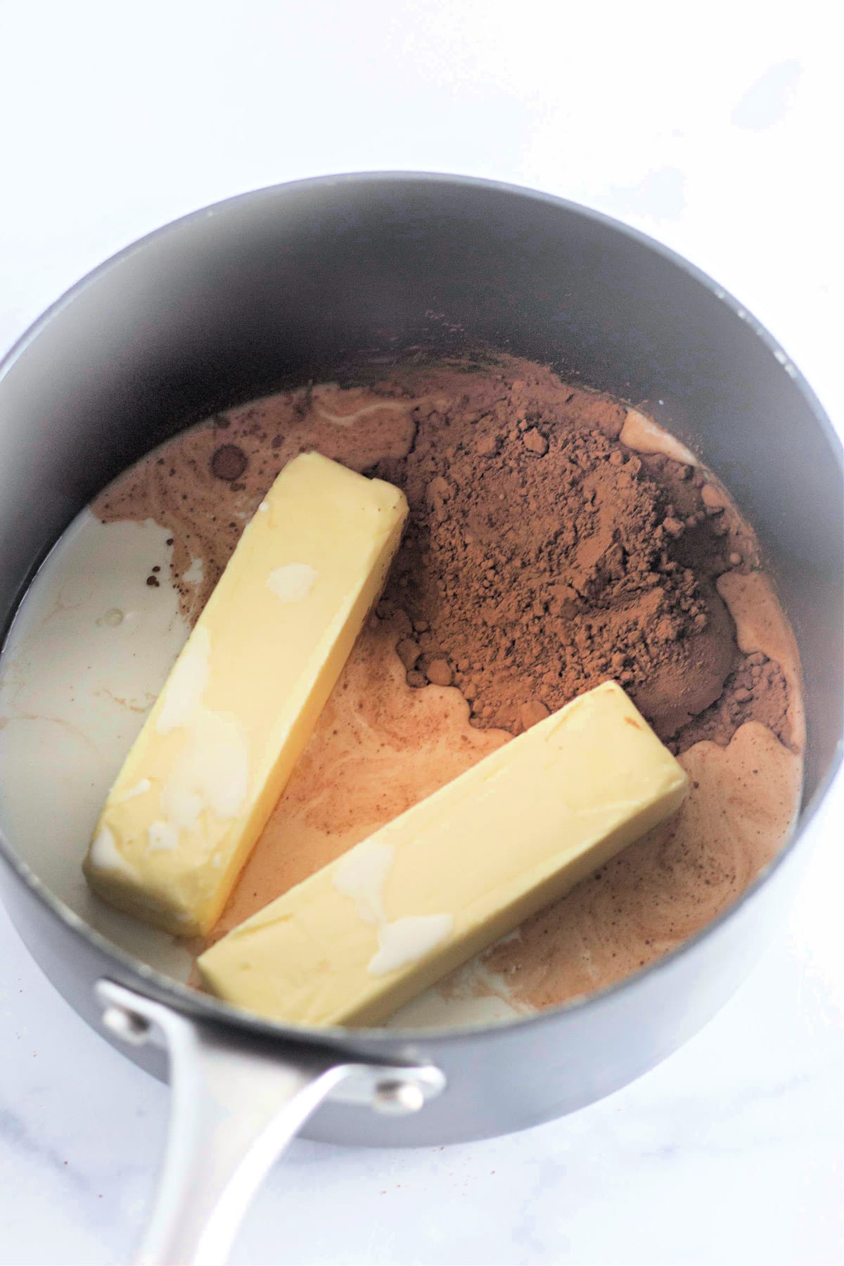 Two sticks of butter and cocoa powder in a saucepan on a white countertop.