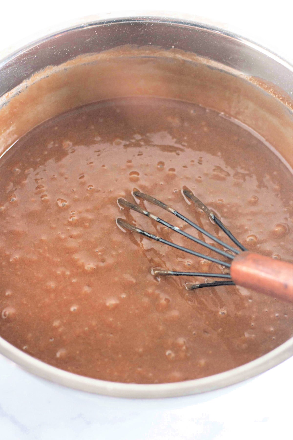 A whisk in a pot of thick brown chocolate batter with bubbles on the surface.