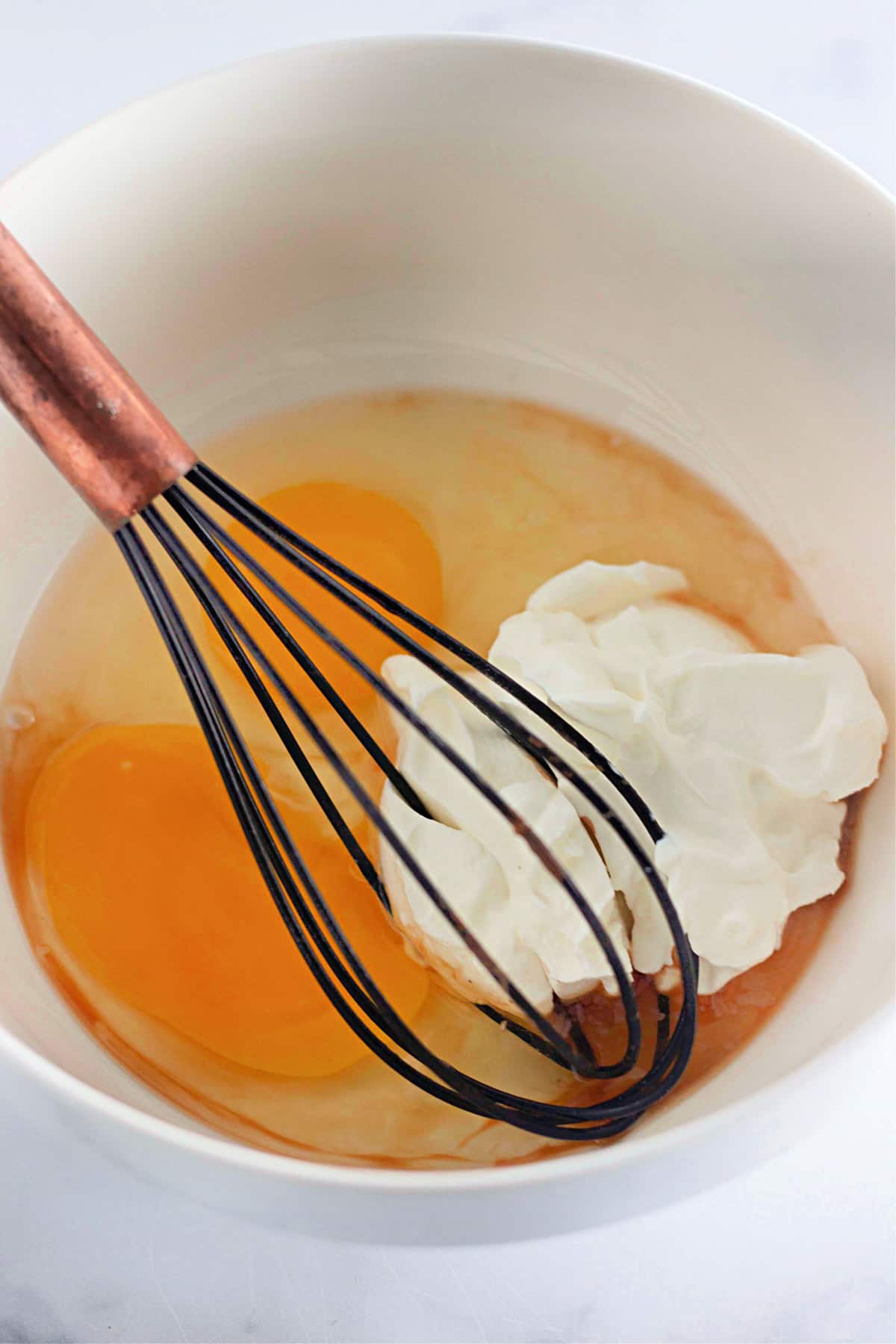 Whisk, eggs, and cream in a white bowl on a marble surface.