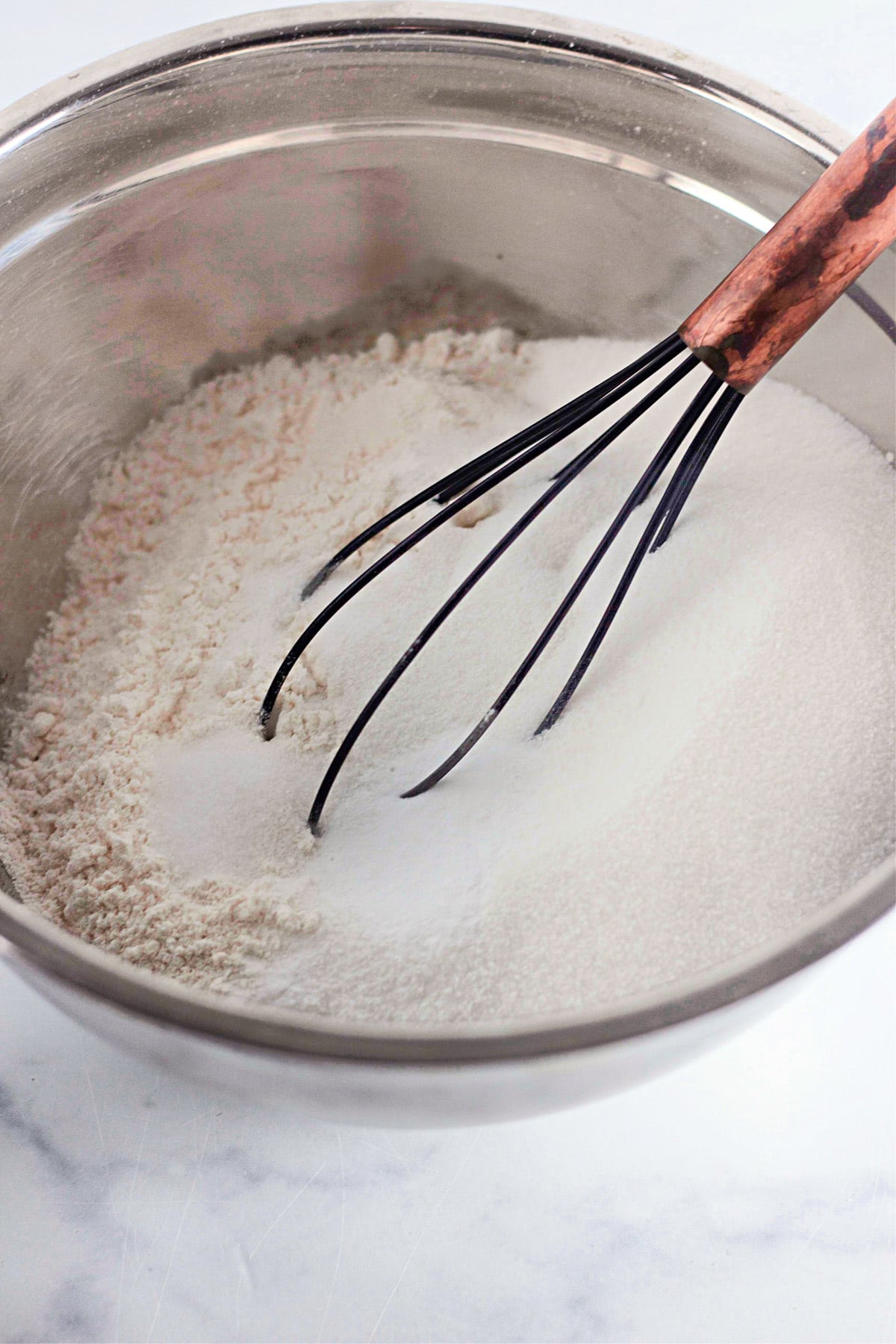 A whisk resting in a metal bowl containing flour and sugar on a white surface.