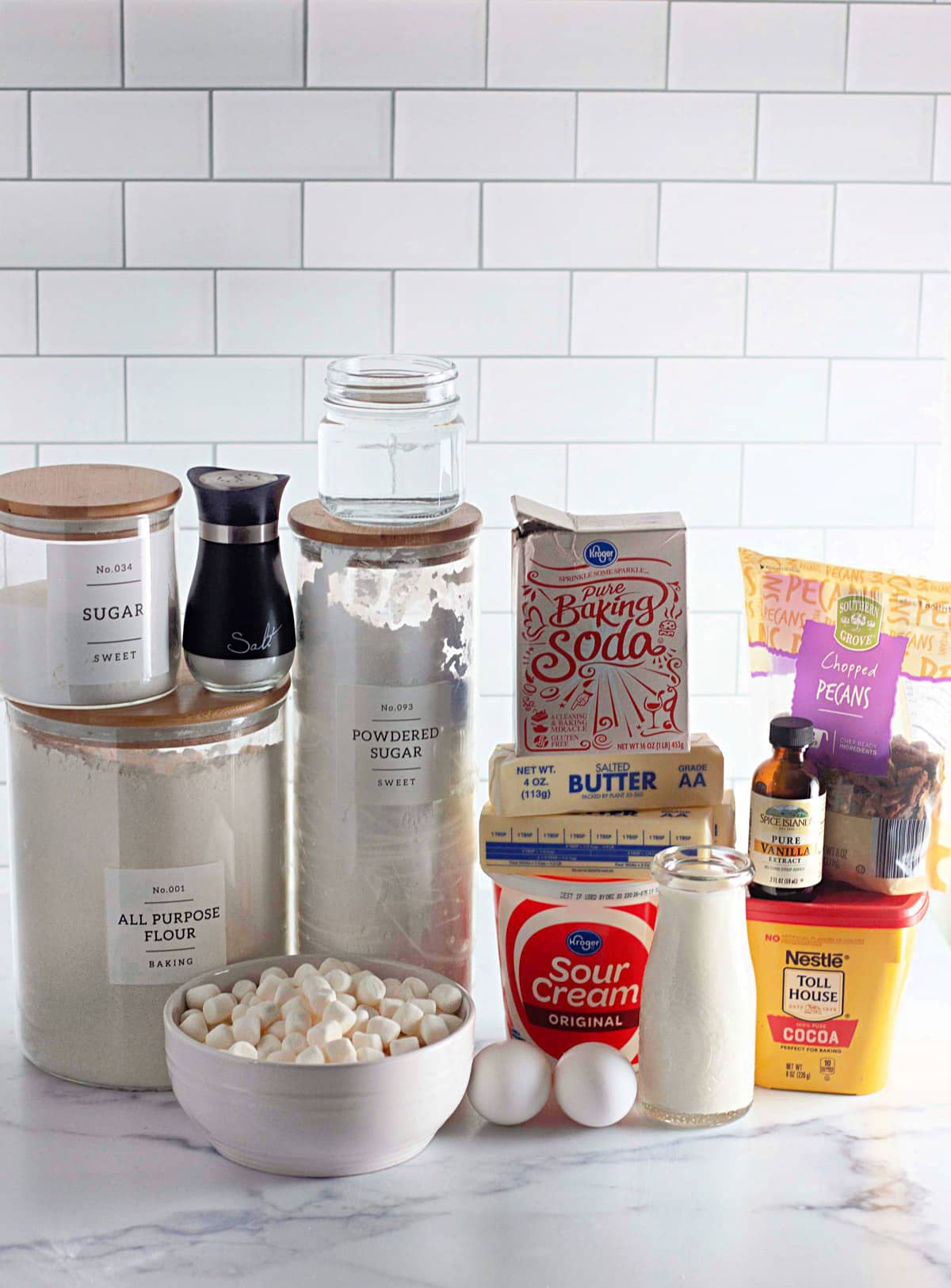 Baking ingredients arranged on a marble counter with canisters labeled for flour, sugar, and more against a tiled wall.