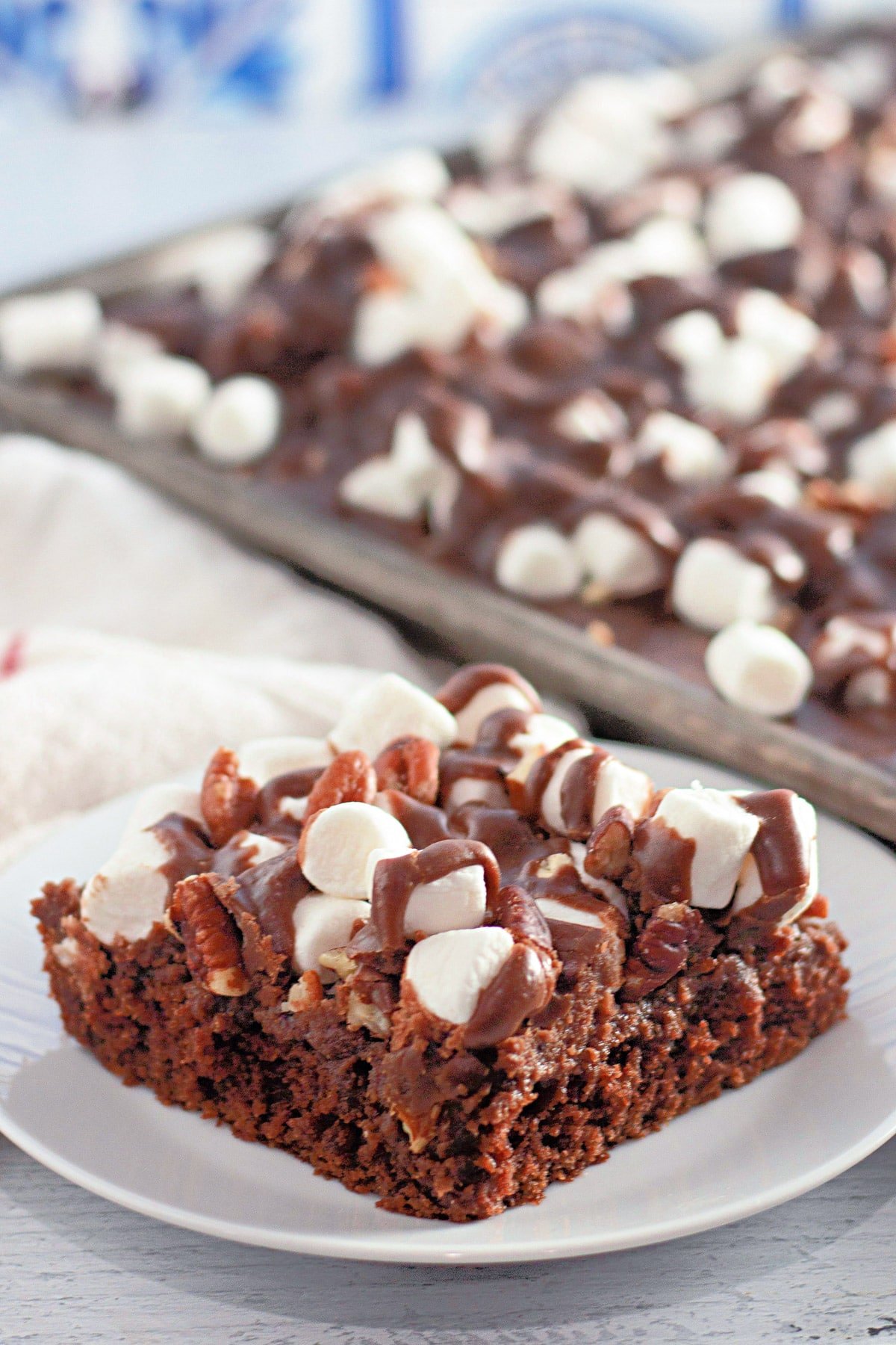 A slice of chocolate cake topped with marshmallows and pecans, drizzled with chocolate sauce, on a white plate.