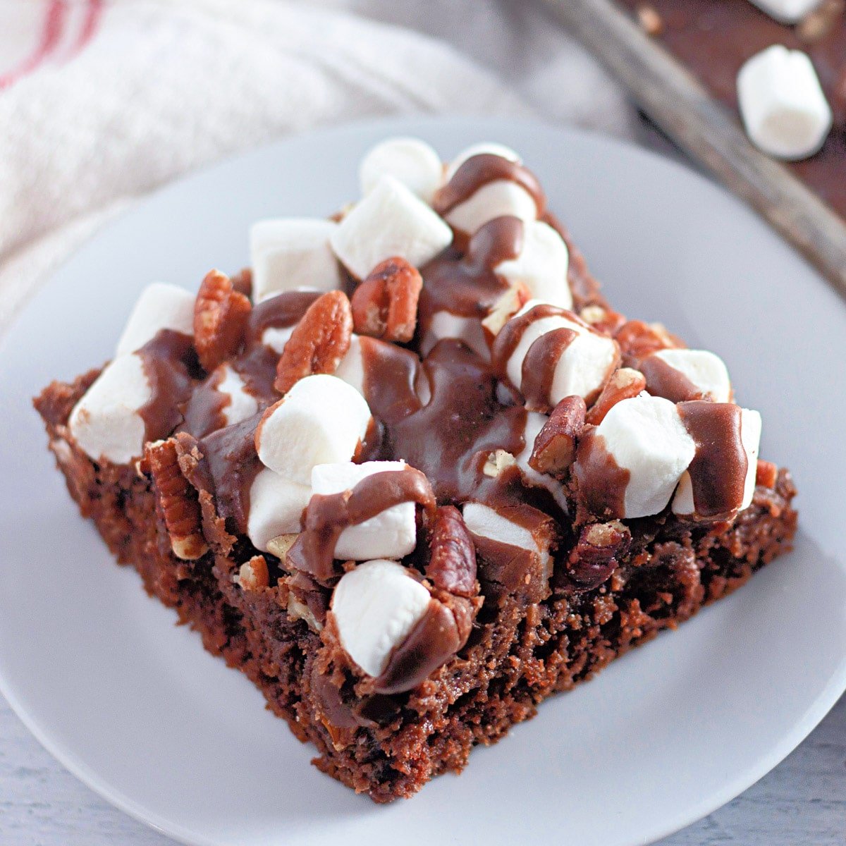 Close-up of a brownie topped with melted chocolate, marshmallows, and pecans on a white plate.