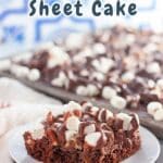 A slice of indulgent Rocky Road Cake, loaded with marshmallows and nuts, sits temptingly on a plate. More of this decadent sheet cake awaits in the background, beckoning for another delicious serving.