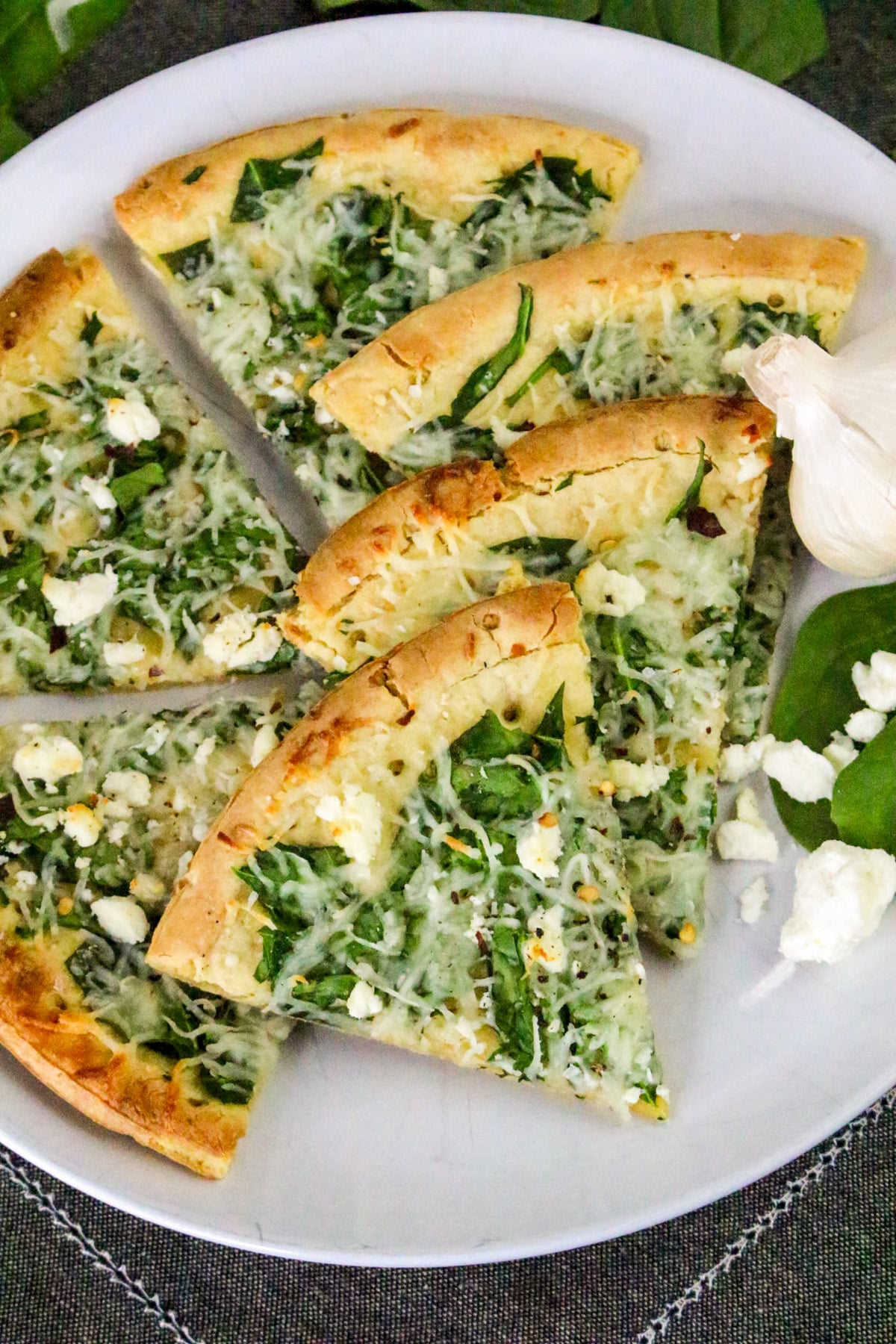 Sliced spinach and cheese pizza on a white plate with garlic and spinach leaves garnish.