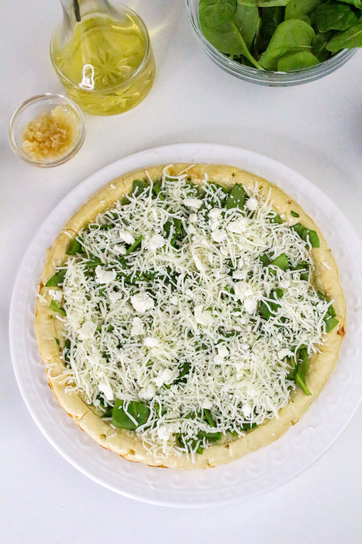 Unbaked pizza with spinach and cheese on a white plate, surrounded by garlic, olive oil, and a bowl of spinach.