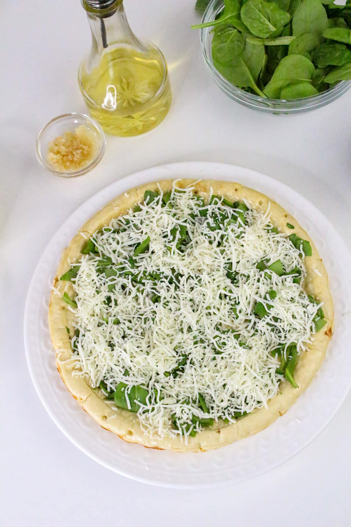Unbaked pizza with cheese and spinach on crust, surrounded by garlic, olive oil, and fresh spinach on a white surface.