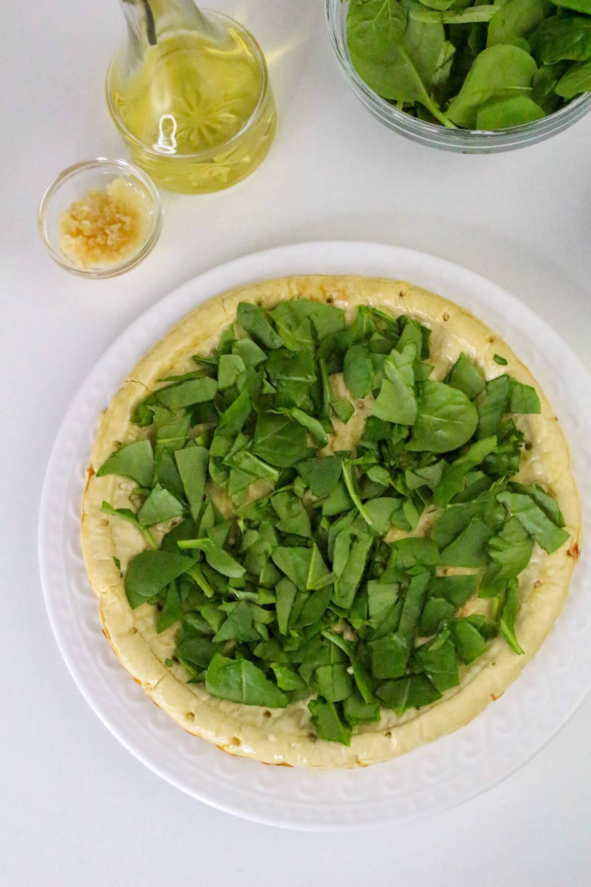 Unbaked pizza with spinach leaves on dough, next to garlic and olive oil on a white surface.