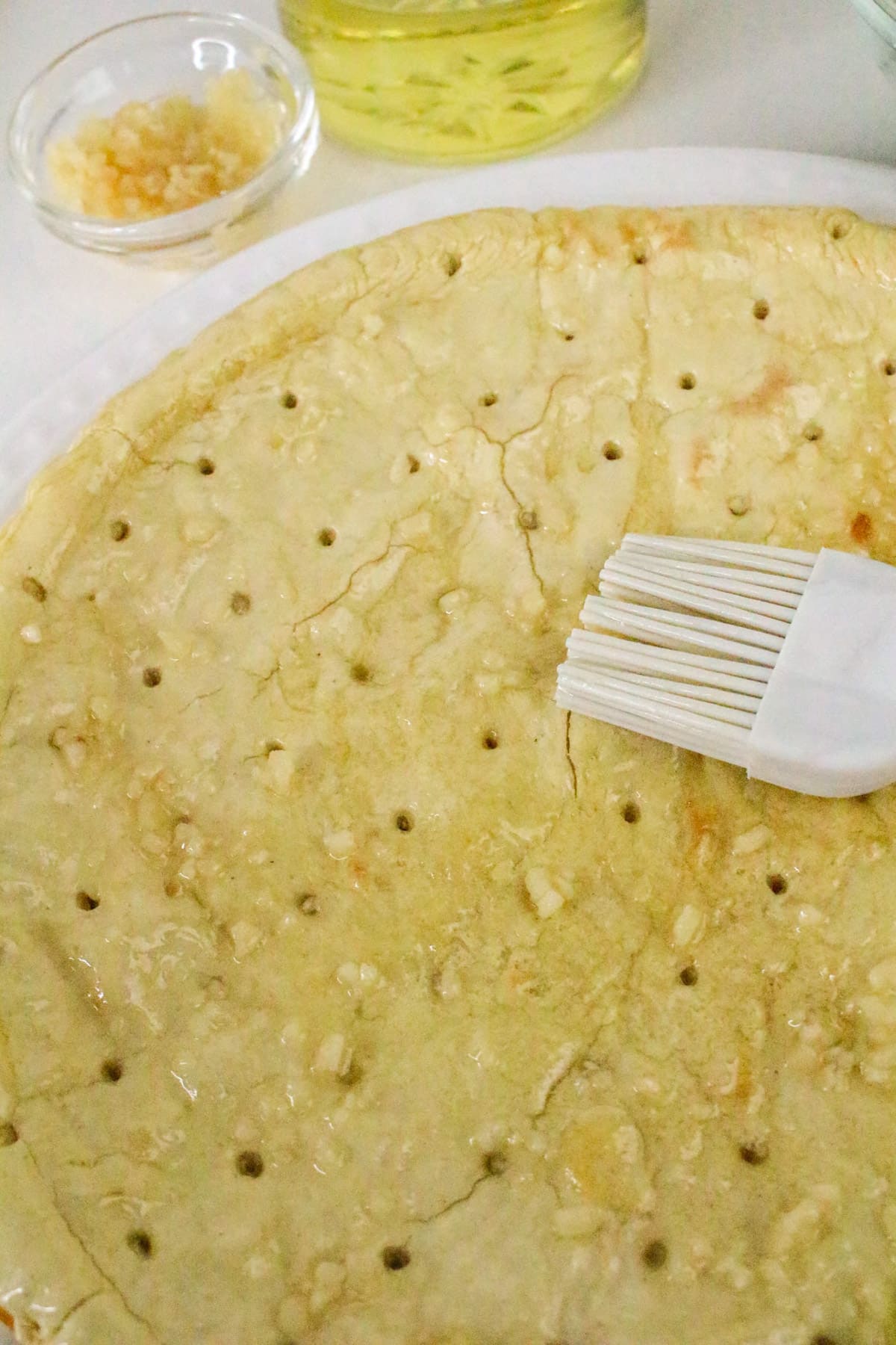 Unbaked pie crust being brushed with garlic oil; minced garlic and oil bowl in the background.