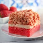 A slice of strawberry crumb cake rests on a white plate, reminiscent of delightful strawberry crunch bars, with fresh strawberries adorning the background.