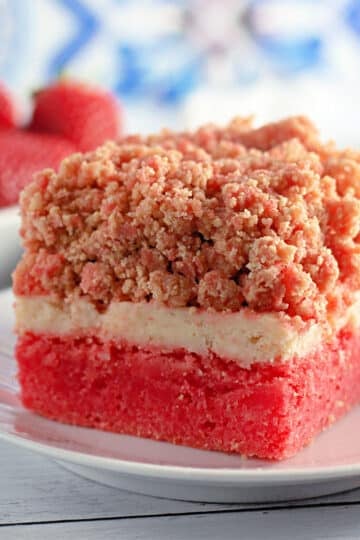 A slice of strawberry crumb cake rests on a white plate, reminiscent of delightful strawberry crunch bars, with fresh strawberries adorning the background.