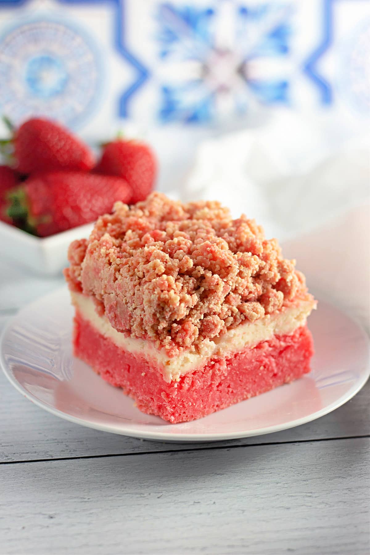A slice of strawberry crumble cake on a white plate, with fresh strawberries and patterned tiles in the background.