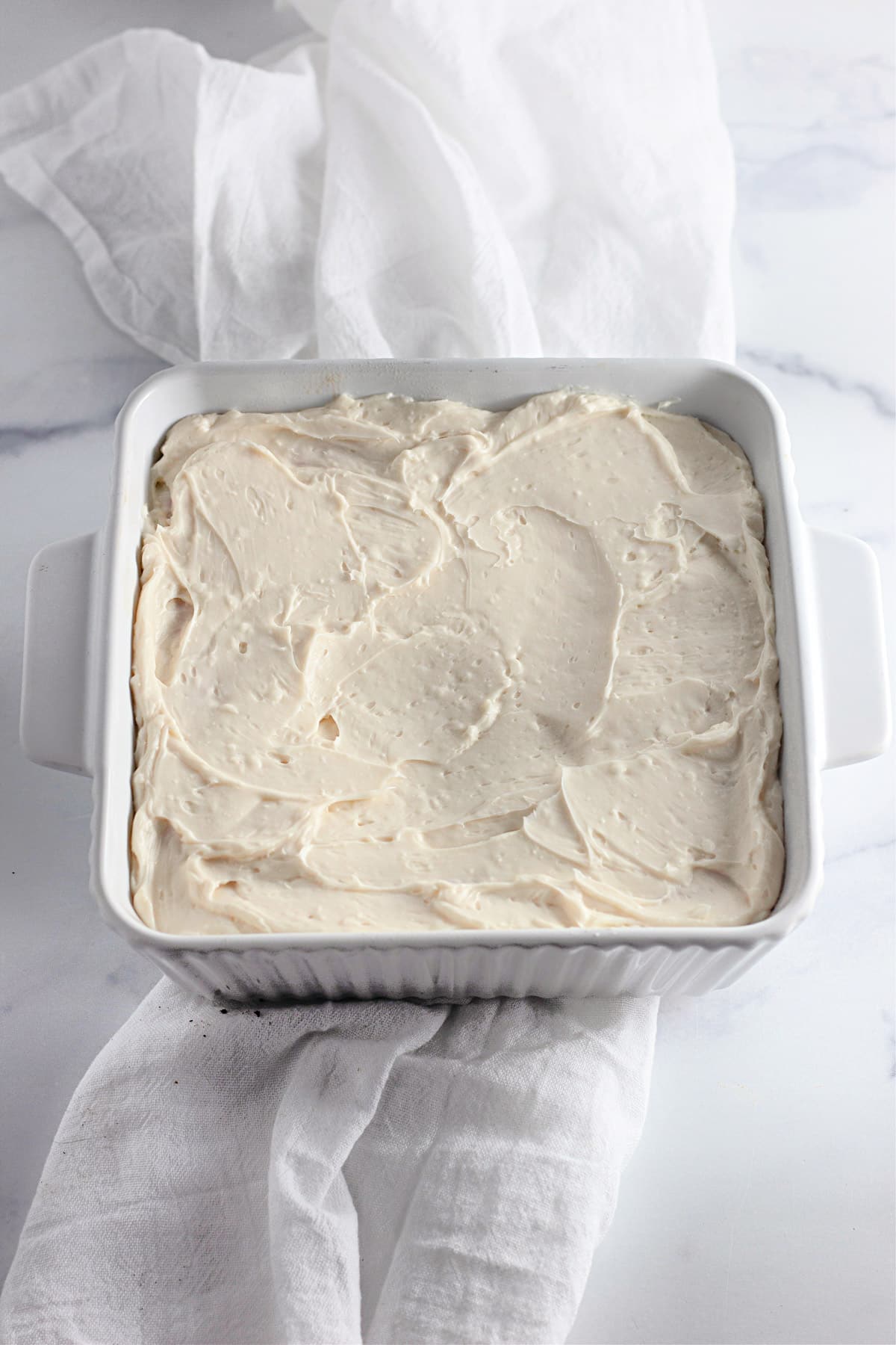 Square white dish with a creamy, textured surface, placed on a white cloth on a marble background.