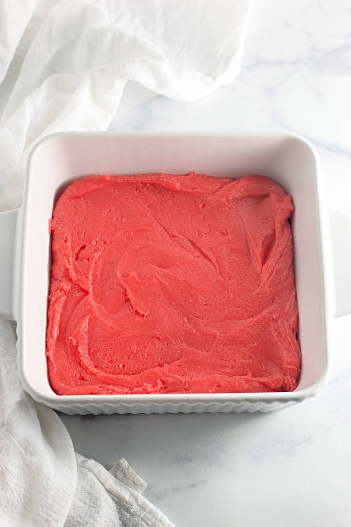 A square dish filled with smooth, pink strawberry cake batter on a white marble surface.