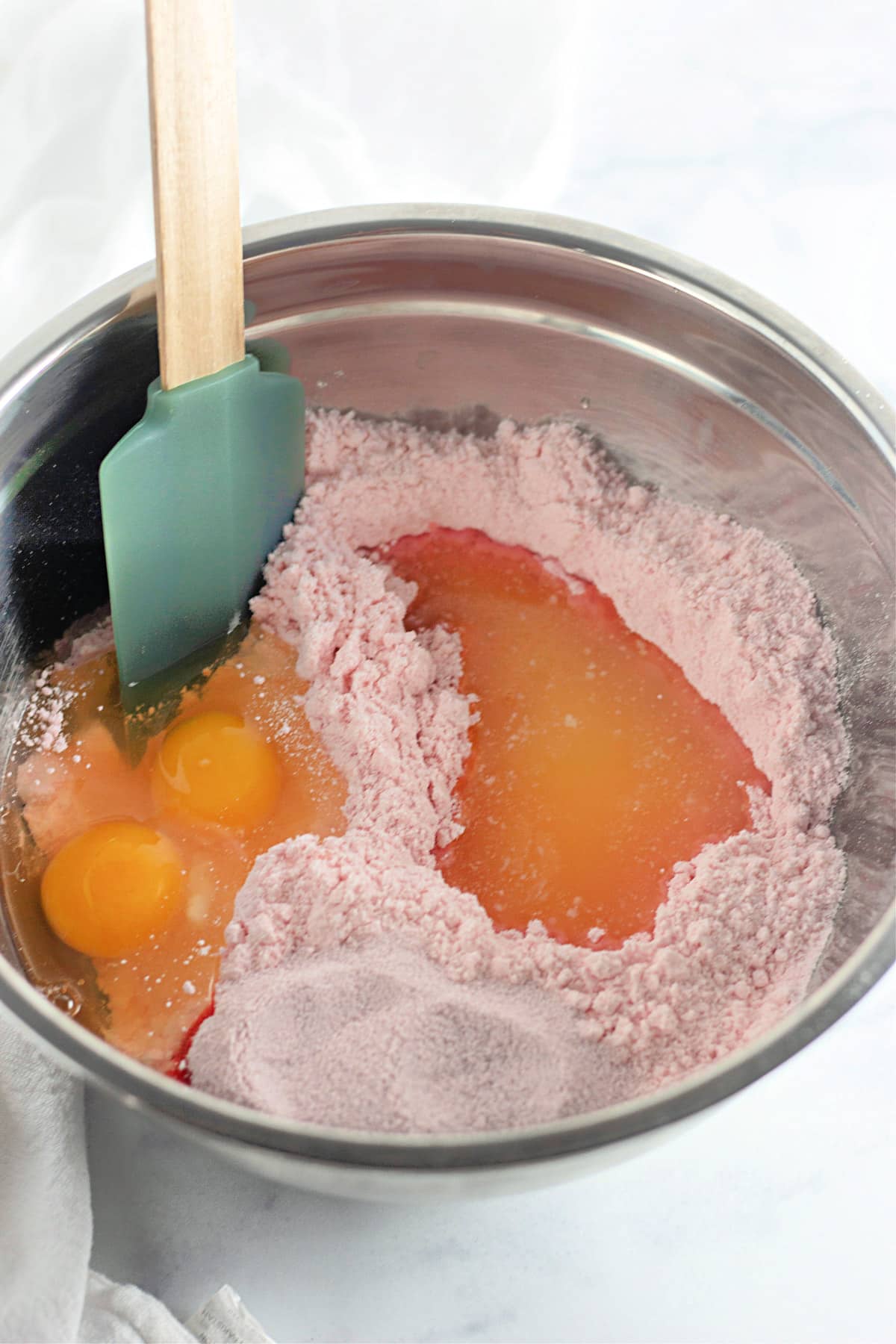 Bowl with pink cake mix, eggs, and water, being mixed with a green spatula.