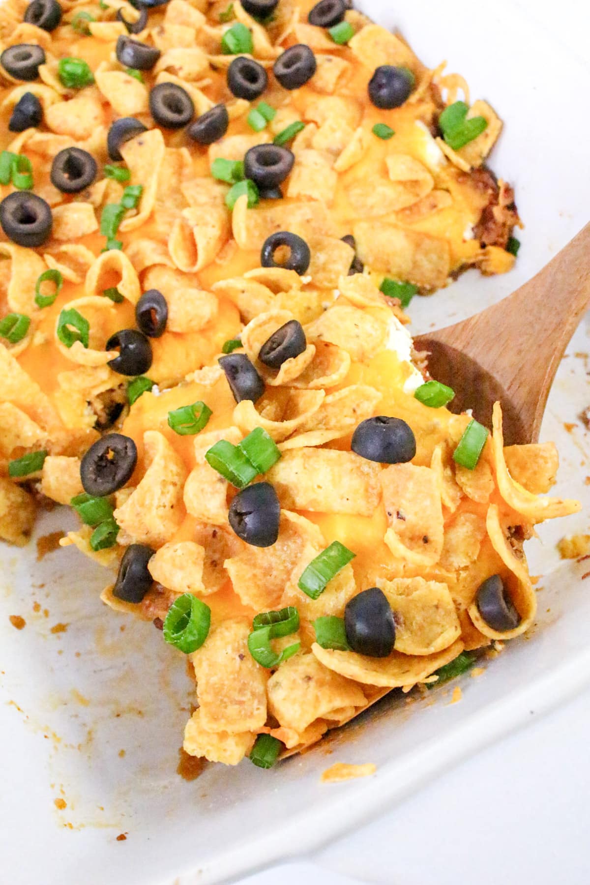 A baked casserole topped with Fritos, melted cheese, sliced black olives, and green onions, served with a wooden spatula.