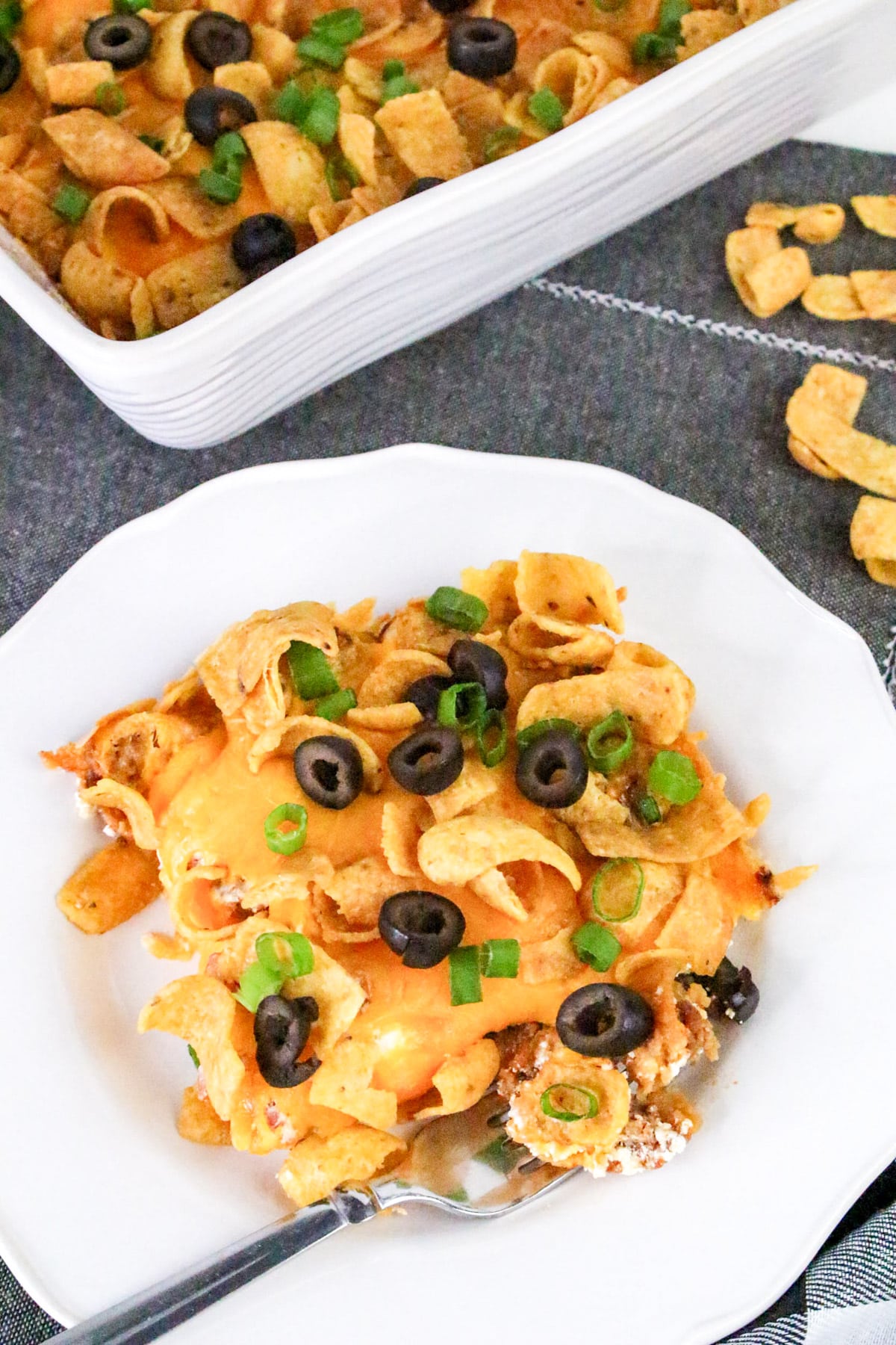 A plate of casserole with Fritos, melted cheese, sliced black olives, and green onions. A serving dish is in the background.