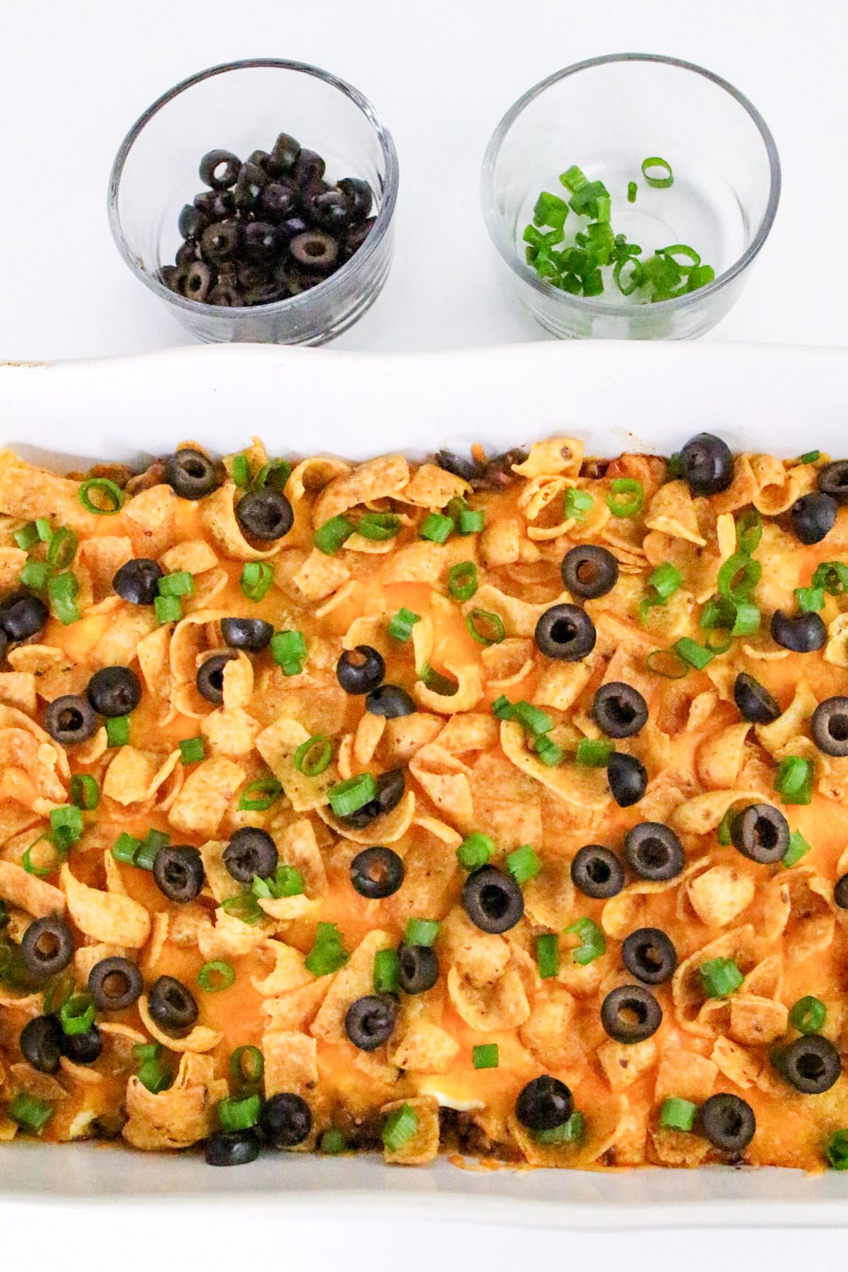 Casserole topped with Fritos, melted cheese, olives, and green onions, served with small bowls of olives and onions.