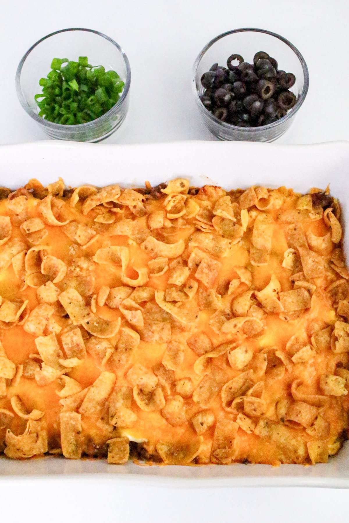 Casserole topped with corn chips and cheese, next to bowls of sliced green onions and black olives on the side.