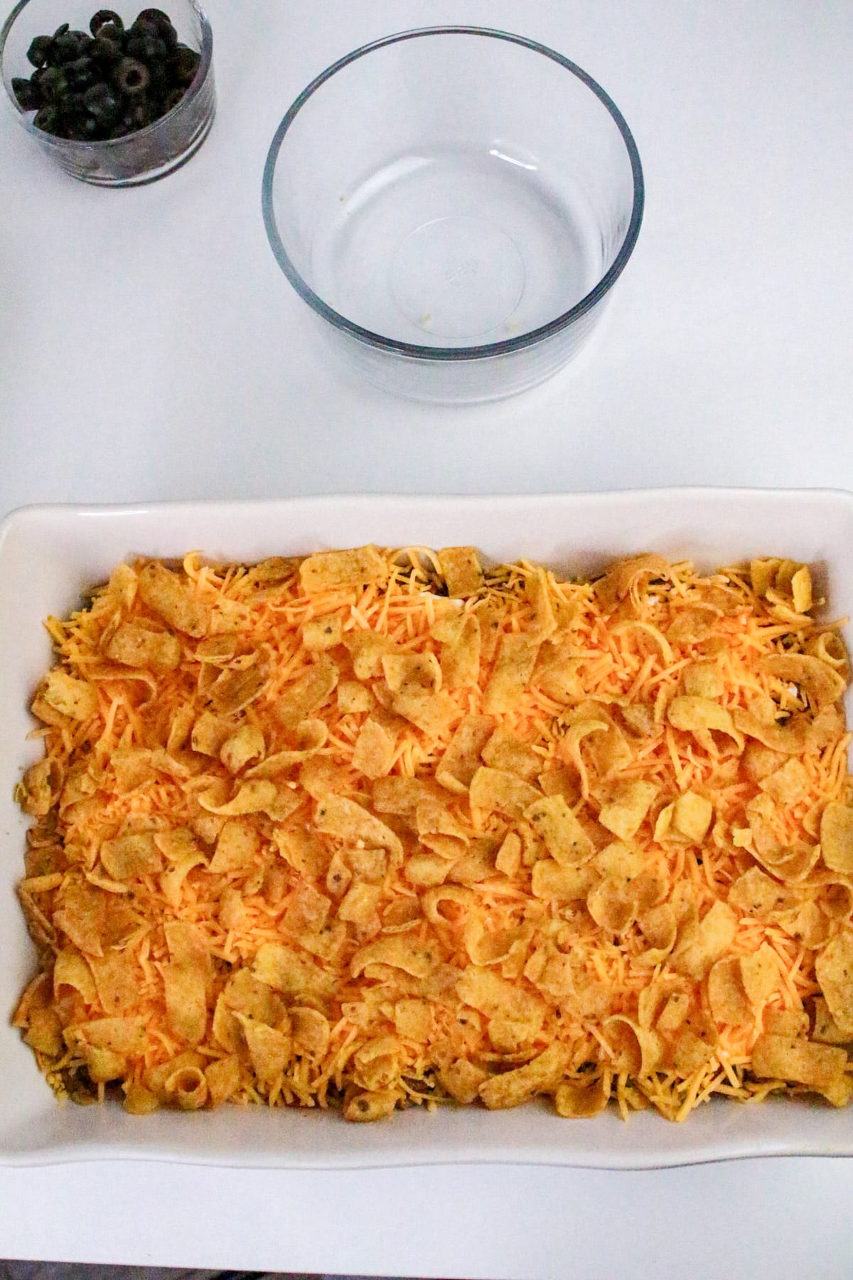 A casserole topped with corn chips and cheese in a white dish; a bowl and sliced olives are nearby.