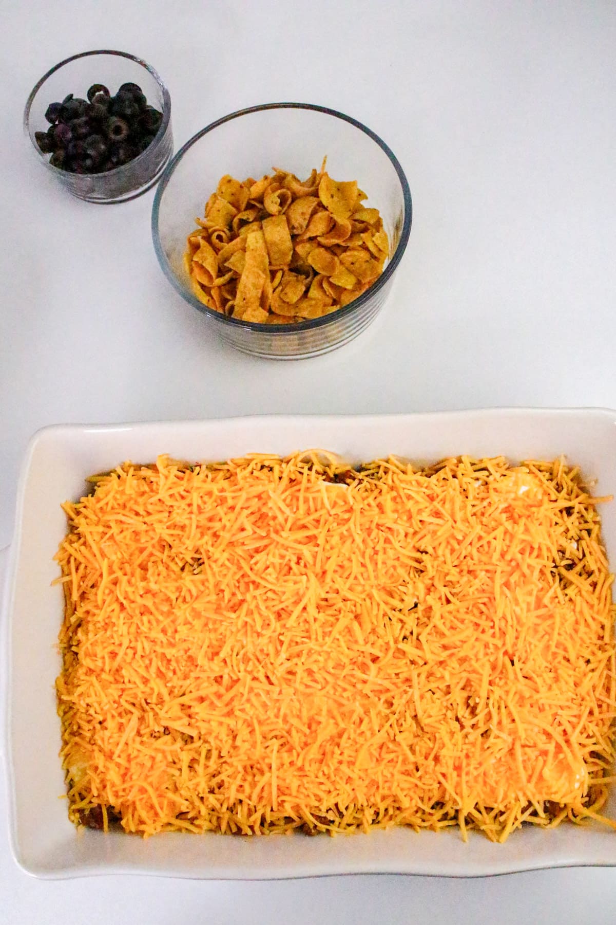 Rectangular dish with cheese-topped casserole, a bowl of tortilla chips, and a bowl of sliced black olives on the side.