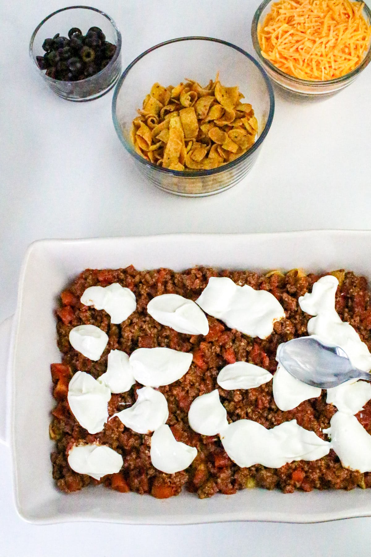 Casserole topped with sour cream in a dish, with bowls of shredded cheese, corn chips, and black olives nearby.