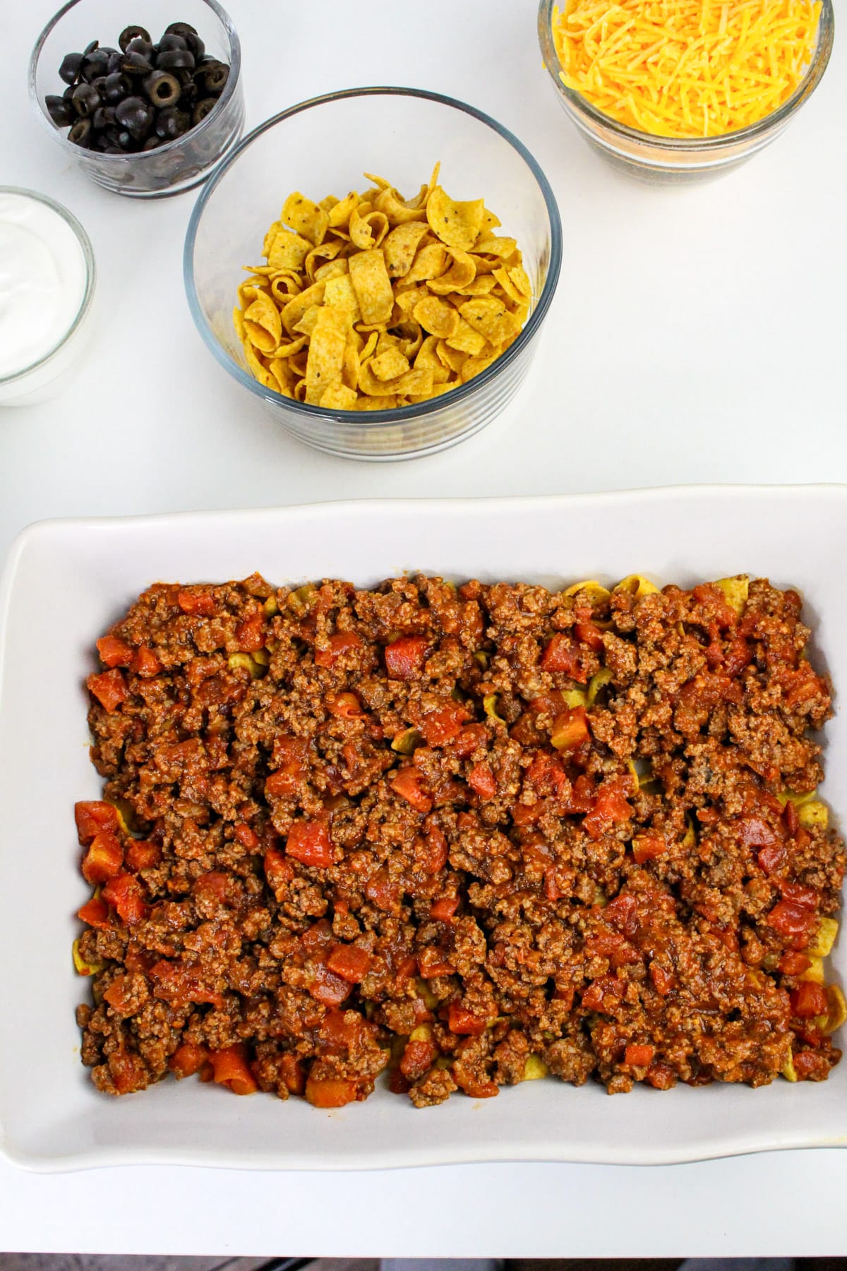 Casserole in a dish with bowls of Fritos, shredded cheese, olives, and sour cream on the side.