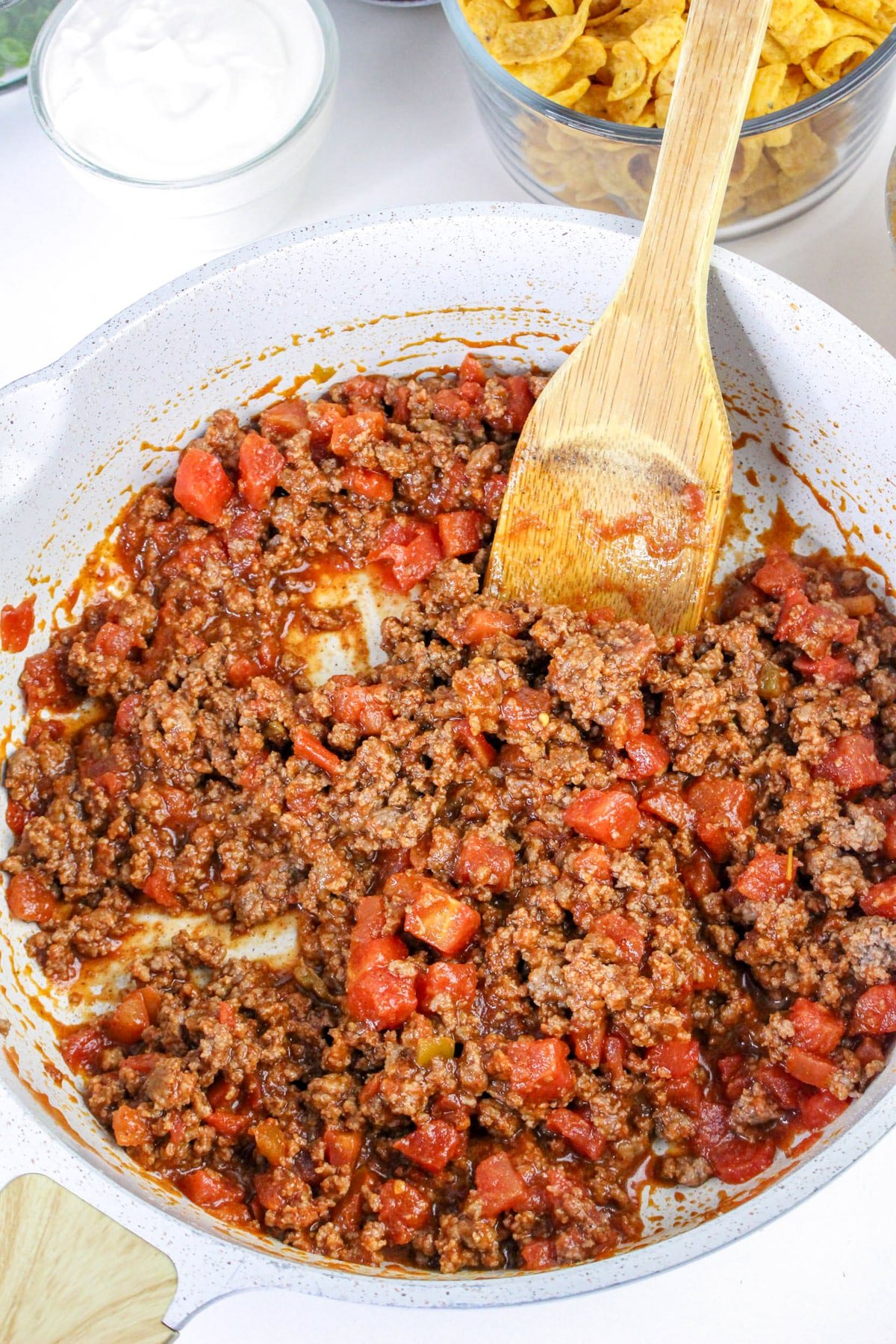 A pan with seasoned ground beef and diced tomatoes, stirred with a wooden spoon. Sour cream and chips in the background.