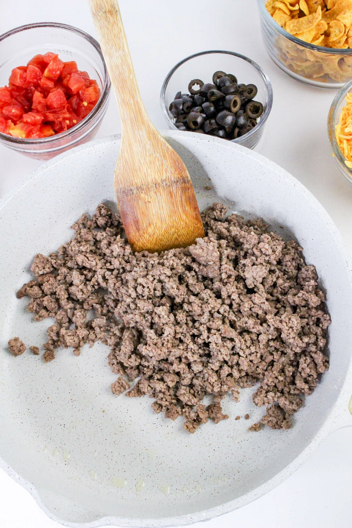 Ground beef cooking in a pan with a wooden spoon, surrounded by bowls of tomatoes, olives, and chips.