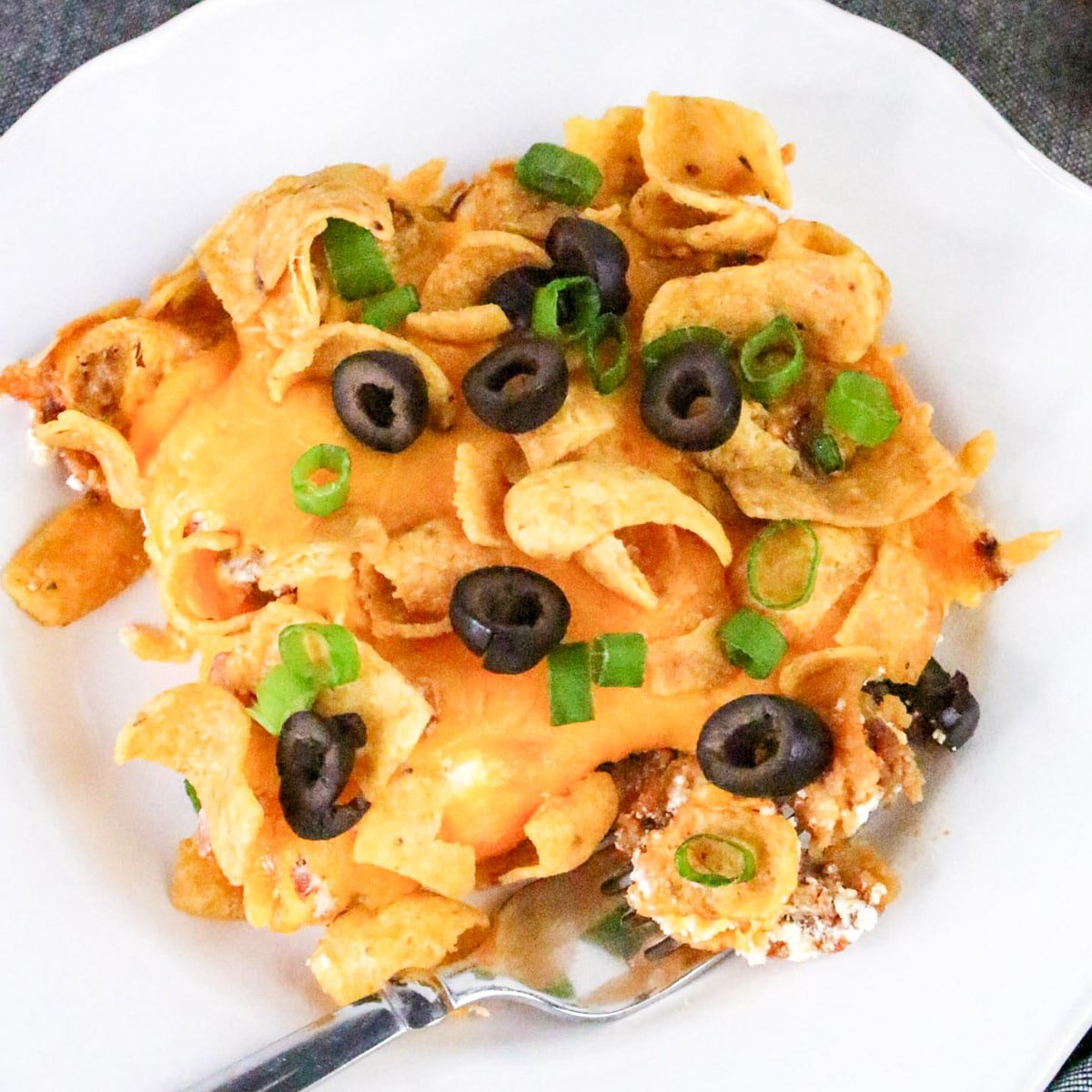 Cheesy casserole topped with Fritos, sliced olives, and green onions on a white plate with a fork.