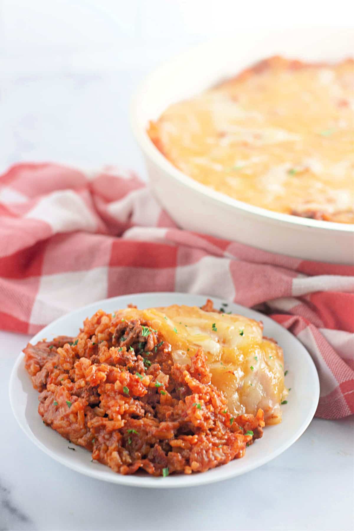 Plate of cheesy, tomato rice casserole with a checkered cloth and a larger casserole dish in the background.