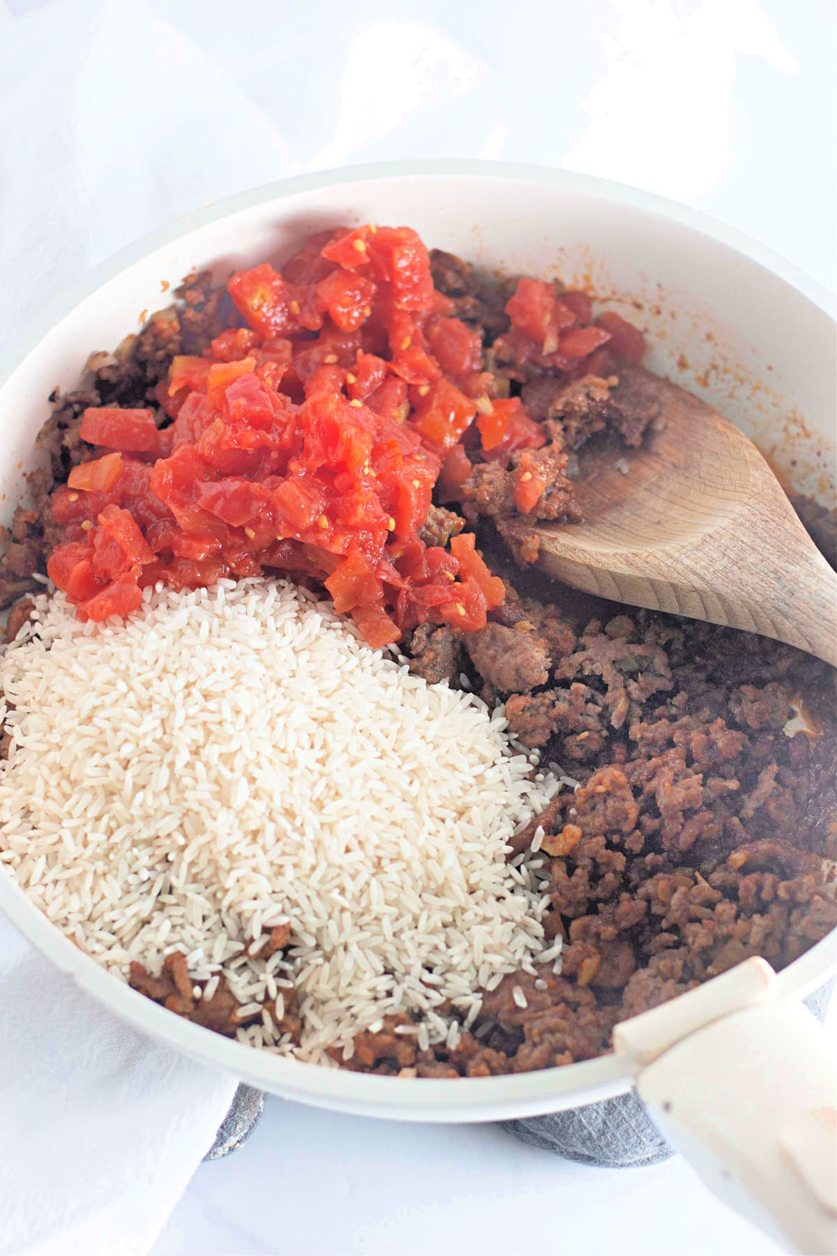 Pan with ground meat, diced tomatoes, and rice, stirred with a wooden spoon.