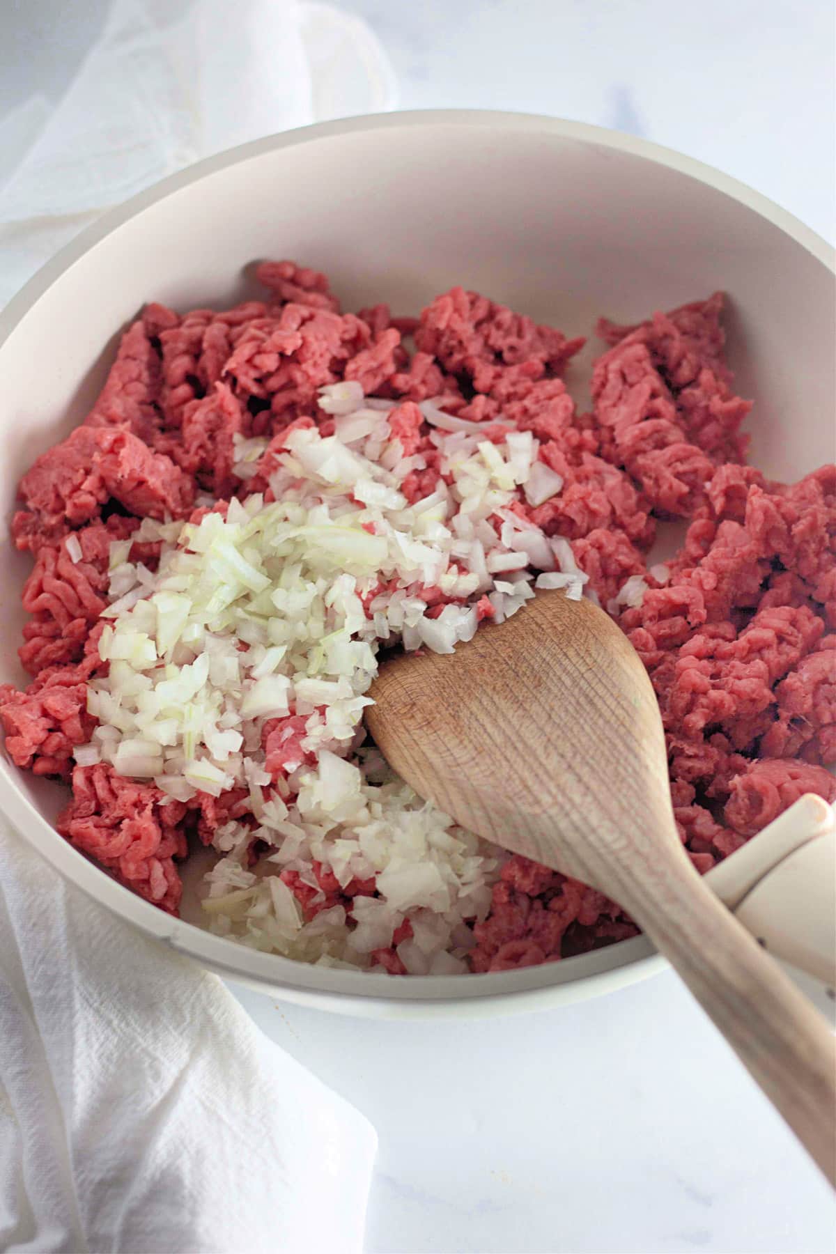 Ground beef and chopped onions in a white pan with a wooden spatula on a white surface.