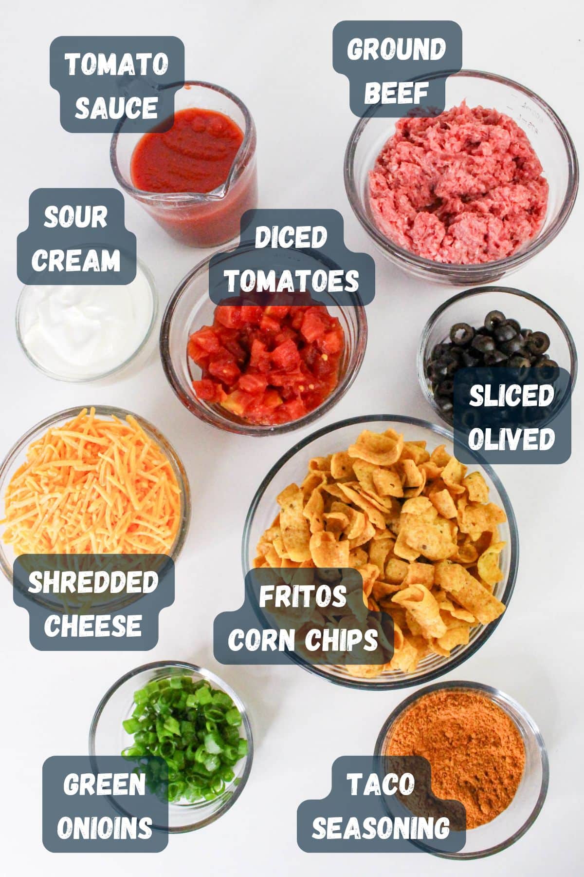 Ingredients in bowls: tomato sauce, ground beef, sour cream, diced tomatoes, sliced olives, cheese, corn chips, onions, seasoning.