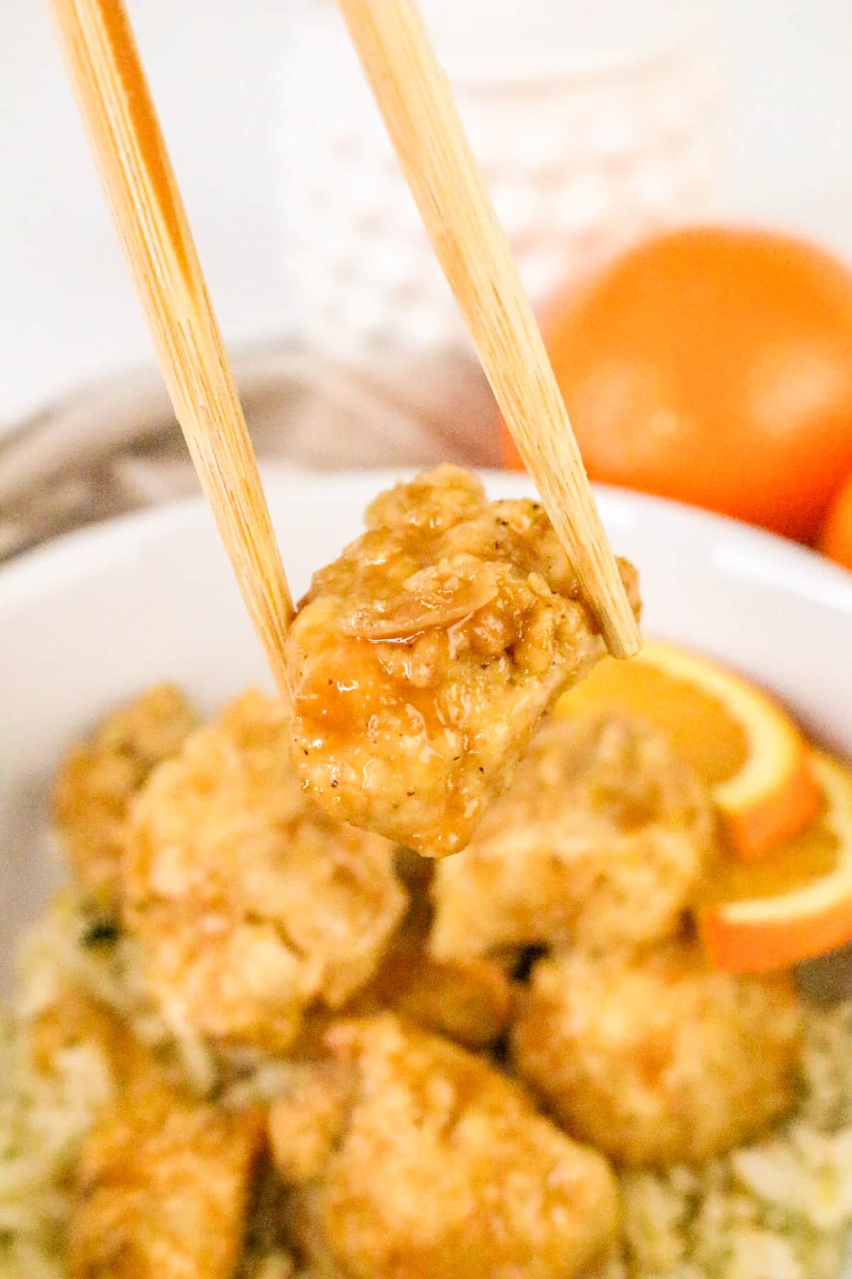 Chopsticks holding a piece of orange chicken above a bowl of rice, garnished with orange slices.