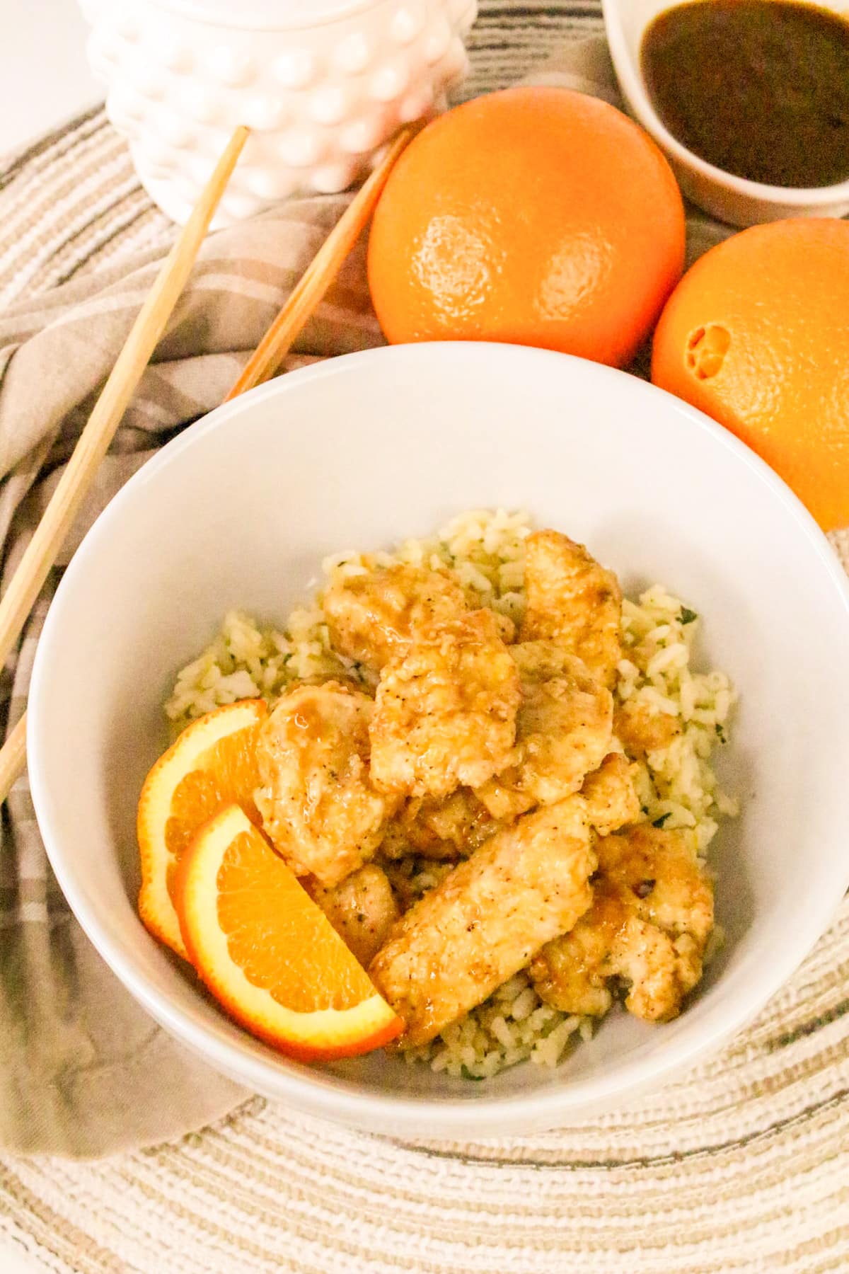 A bowl of orange chicken on rice garnished with orange slices, next to whole oranges and sauce on a textured mat.