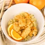 Bowl of orange chicken on rice, garnished with orange slices, with oranges and chopsticks in the background.