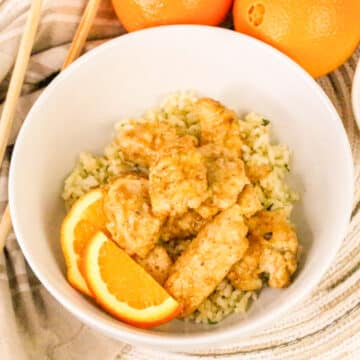 Bowl of orange chicken on rice, garnished with orange slices, with oranges and chopsticks in the background.