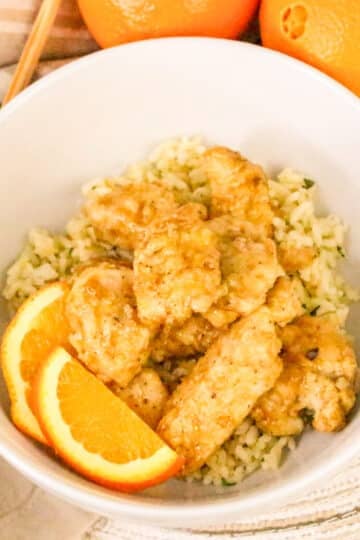 Bowl of orange chicken on rice, garnished with orange slices, with oranges and chopsticks in the background.