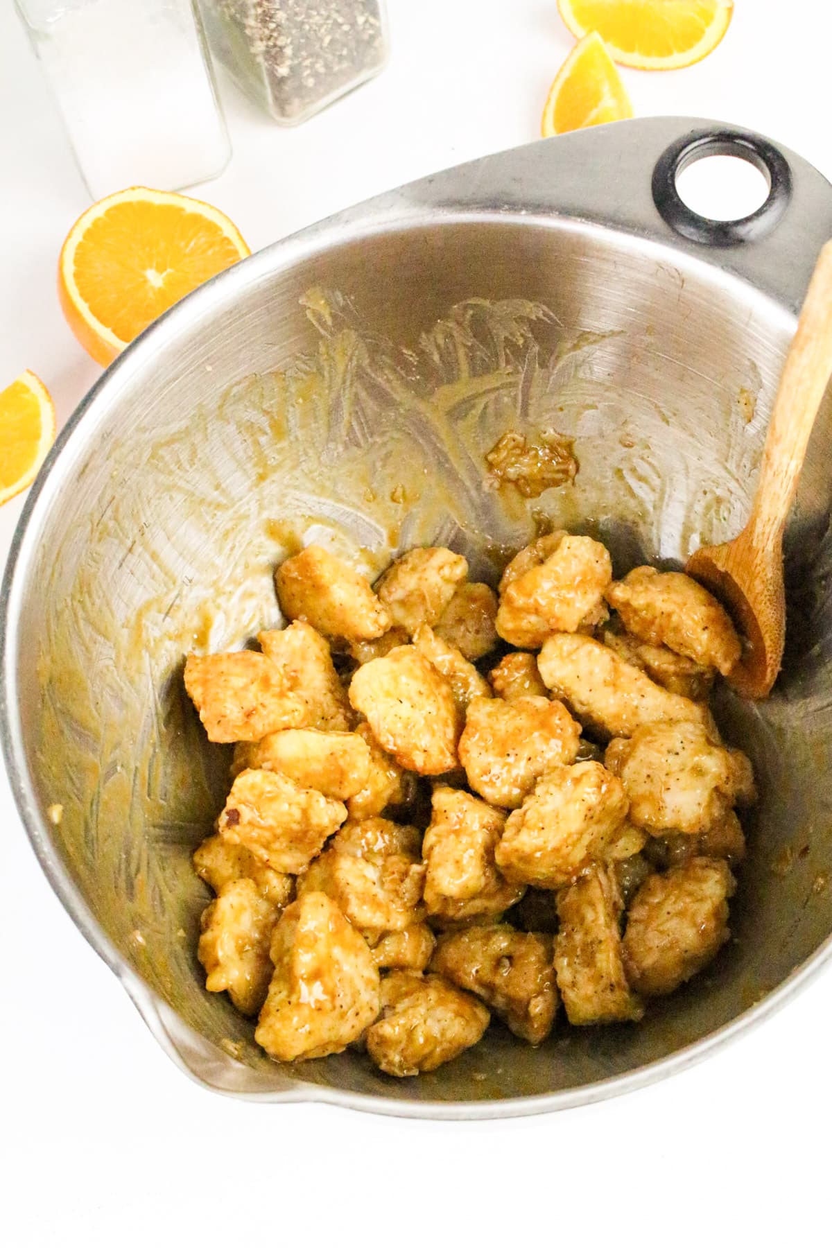 Chicken pieces in sauce inside a mixing bowl, surrounded by orange slices and a wooden spoon.