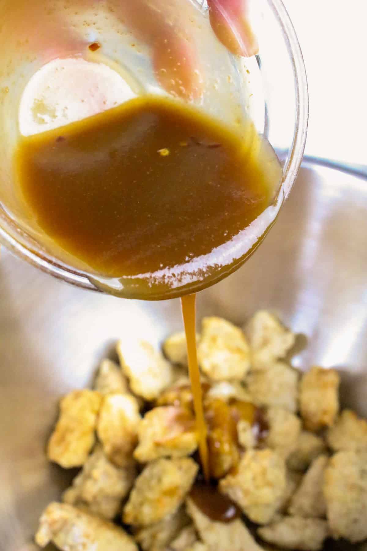 Pouring orange sauce over cooked chicken pieces in a metal bowl.